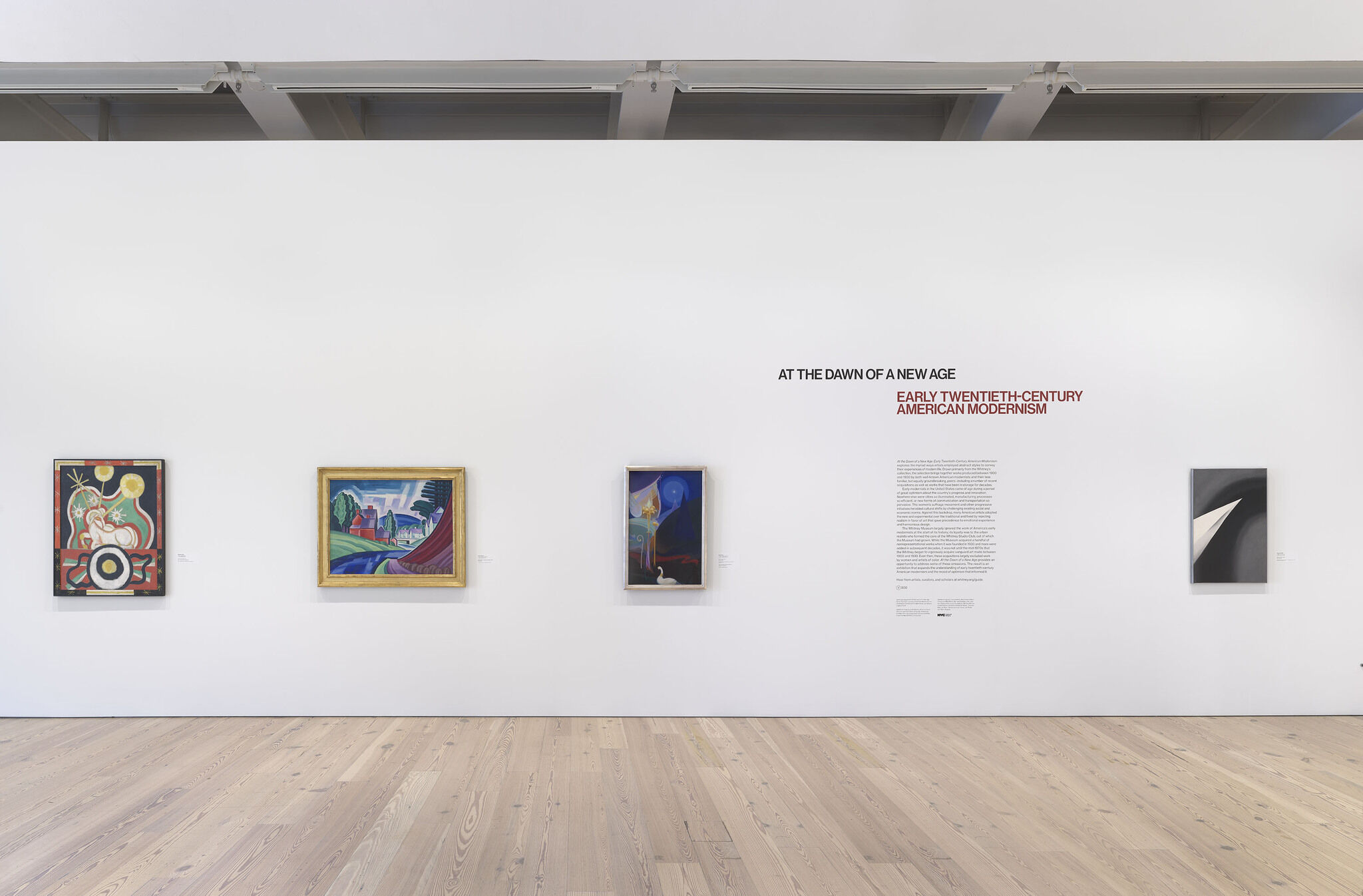 A white exhibition wall with the opening text for At the Dawn of a New Age and four framed works.