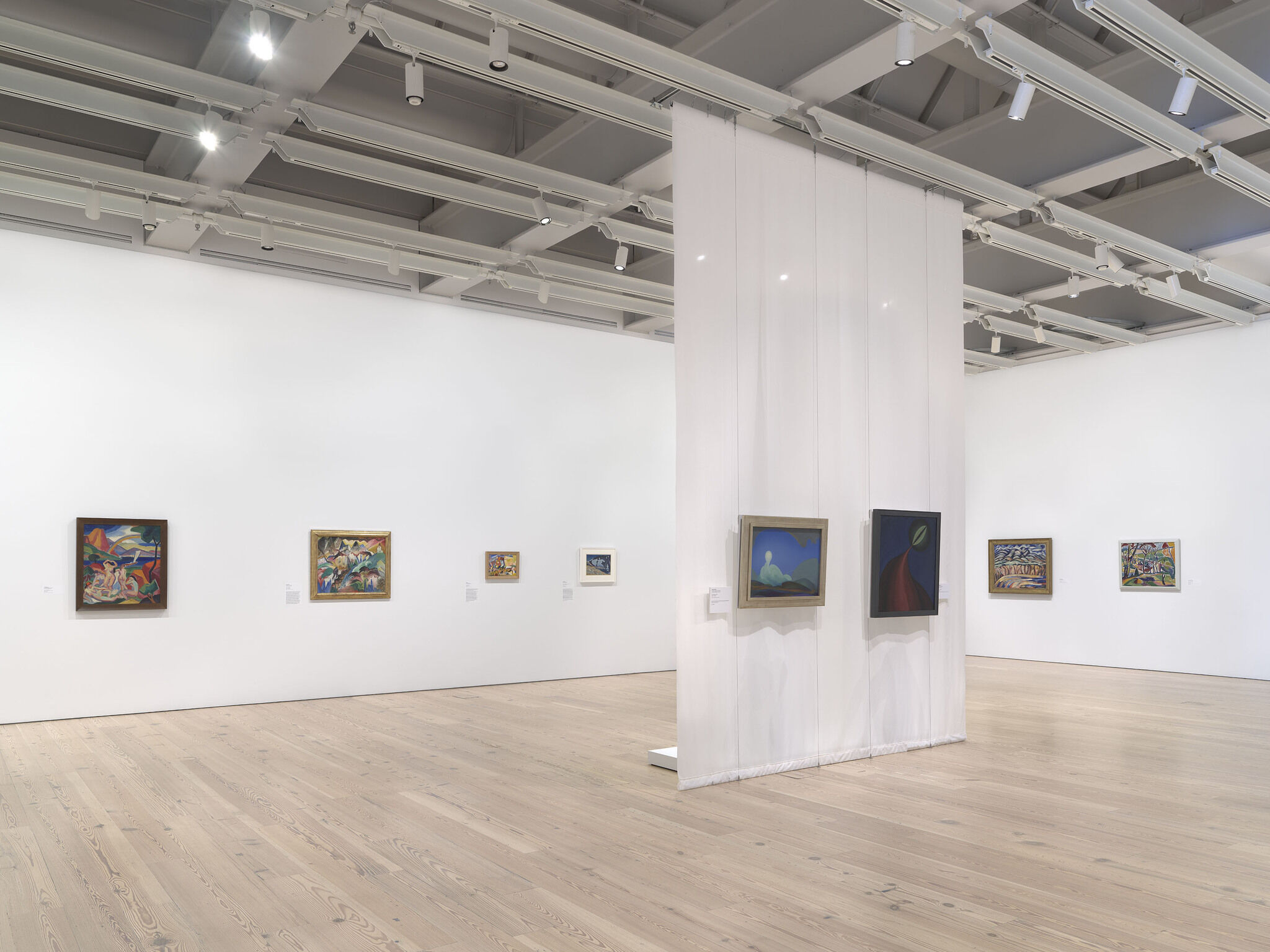 A white exhibition room with six works installed across the outer wall and two installed in the center of the room.