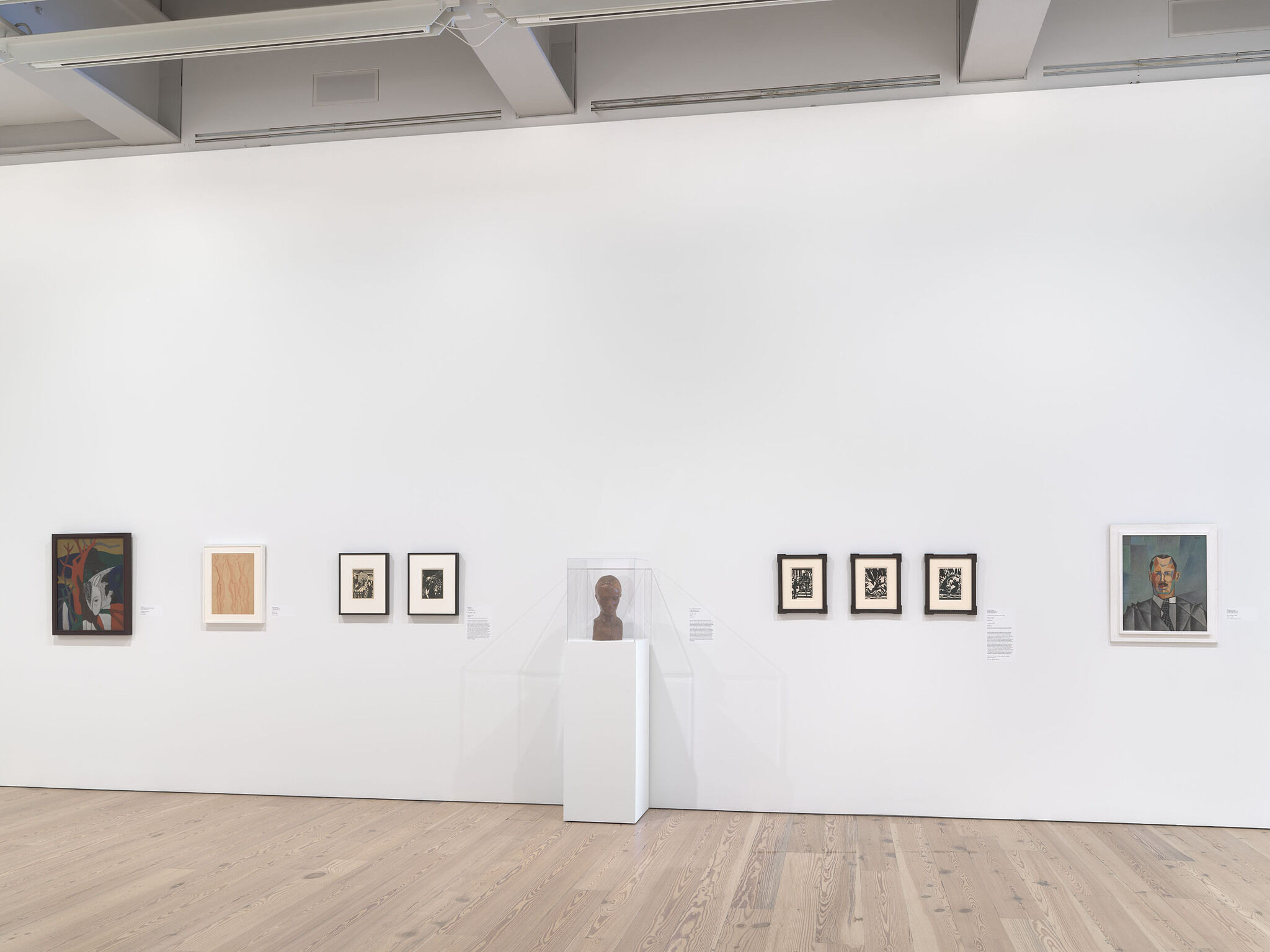 A white exhibition wall with eight works installed across the wall and a ninth work installed in a glass case.