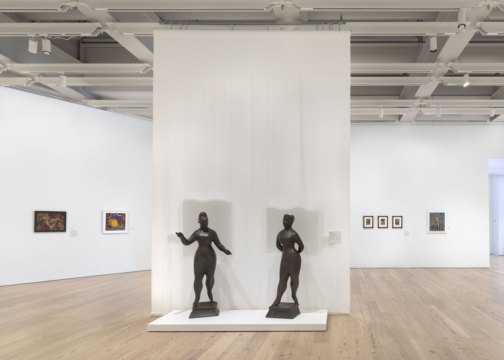 A white exhibition wall with six works installed on the outer wall and two sculptures installed in the center of the room.