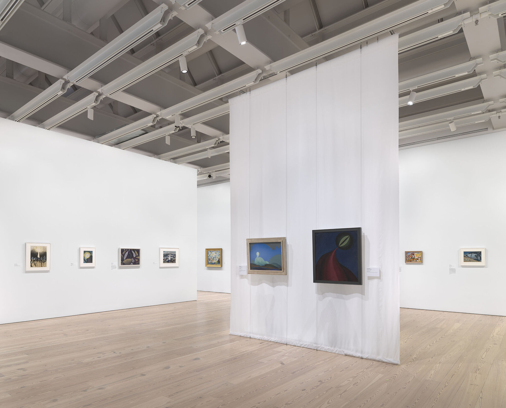 A white exhibition room with seven works installed along the outer wall and two works installed in the center of the room.