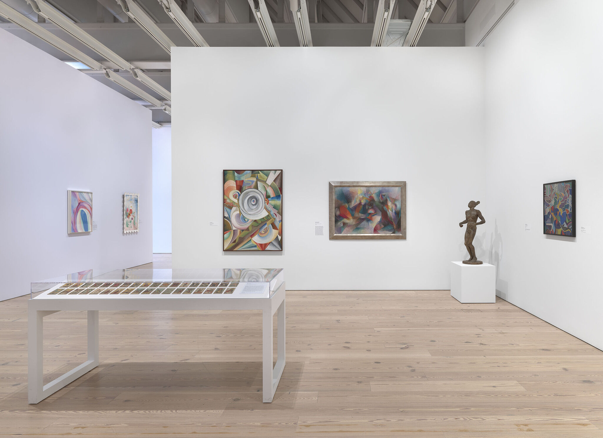 A white exhibition room with five works installed across multiple walls and a work placed in a vitrine in the middle of the room.