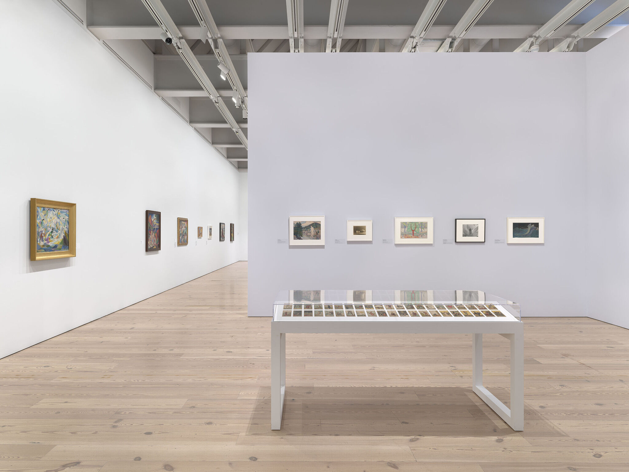 A white exhibition room with twelve works installed across multiple walls and a work on display in a vitrine in the middle of the room.
