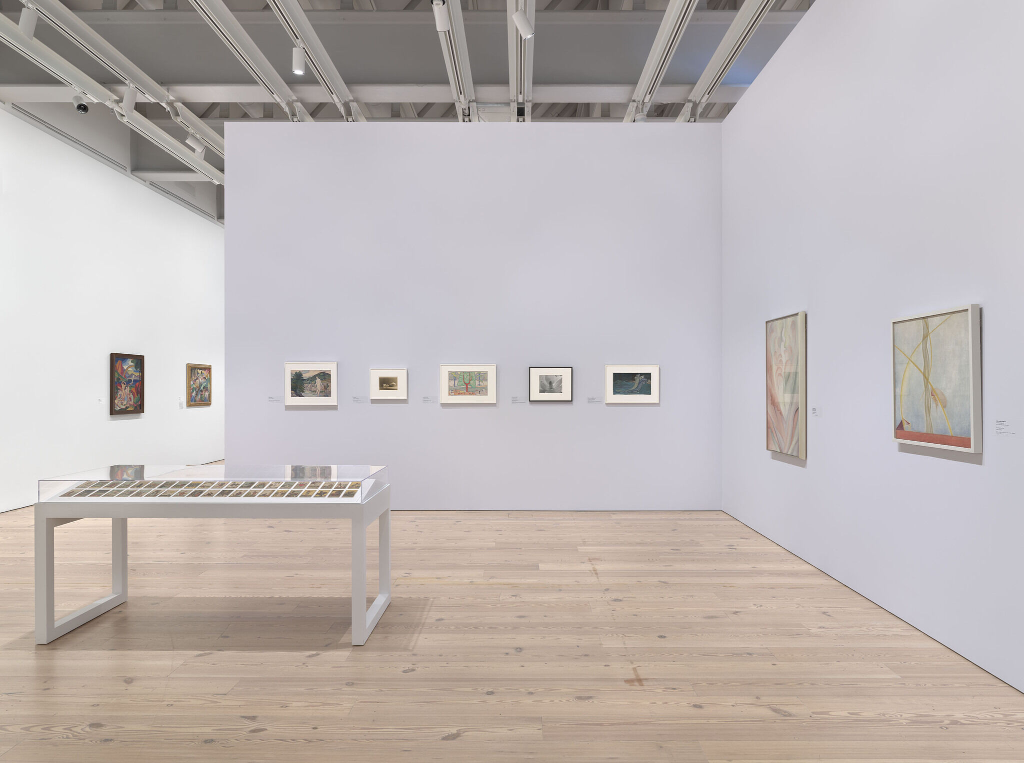 A white exhibition room with nine works installed across multiple walls and a work on display in a vitrine in the middle of the room.