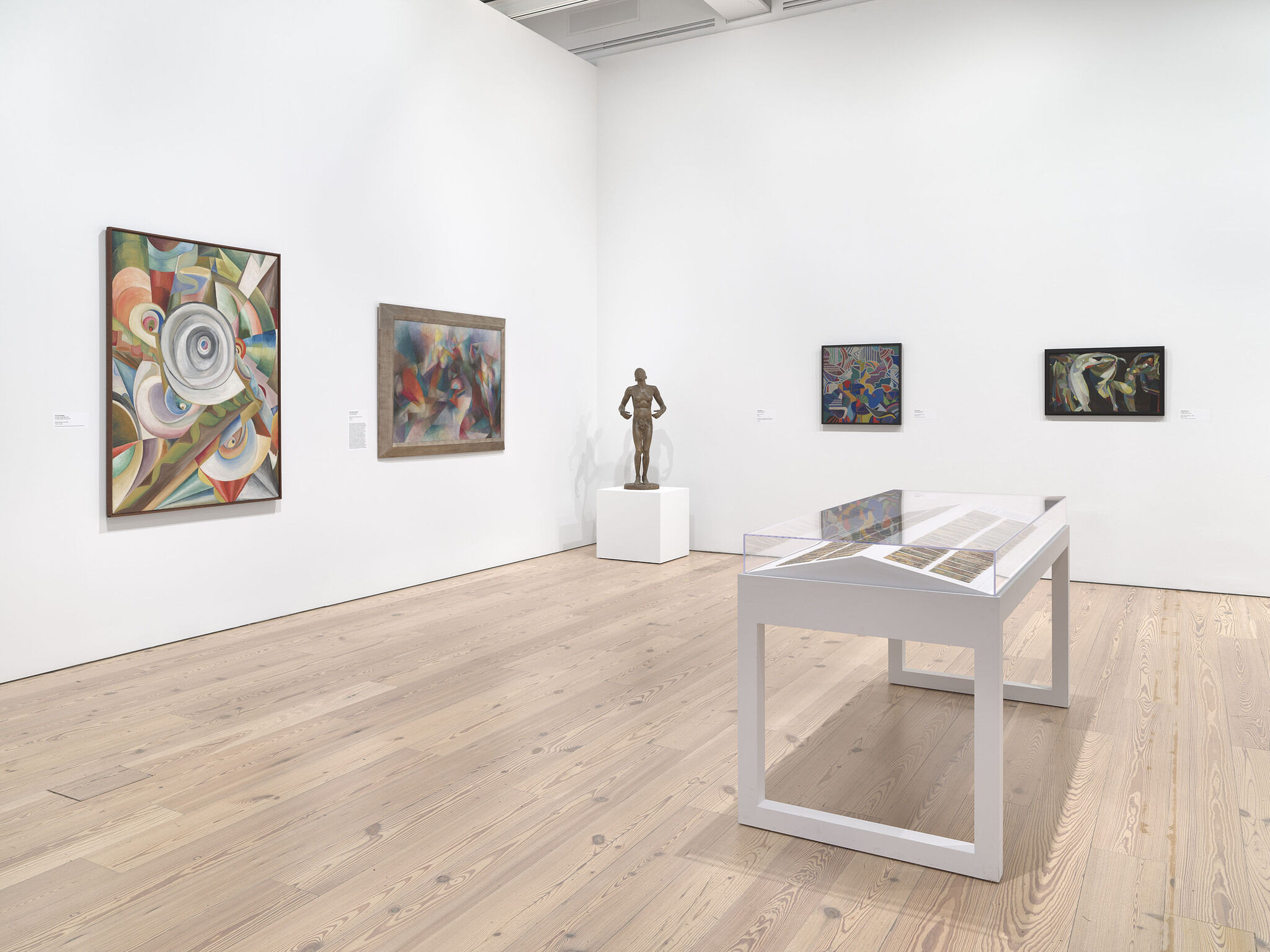 A white exhibition room with four works mounted on the walls and and a fifth work on display in a vitrine in the center of the room.