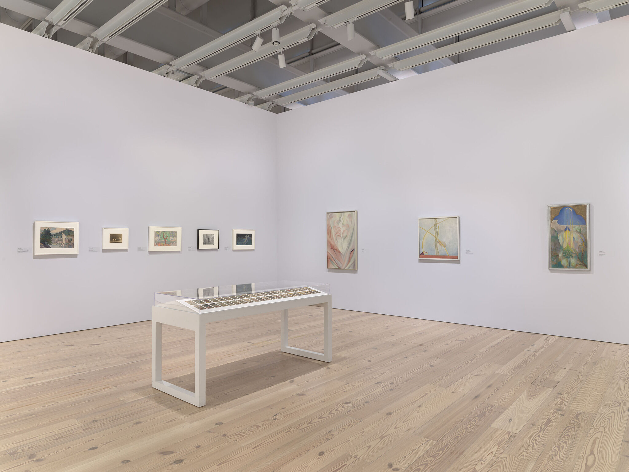 A white exhibition room with eight works mounted on the walls and a ninth on display in a vitrine in the middle of the room.
