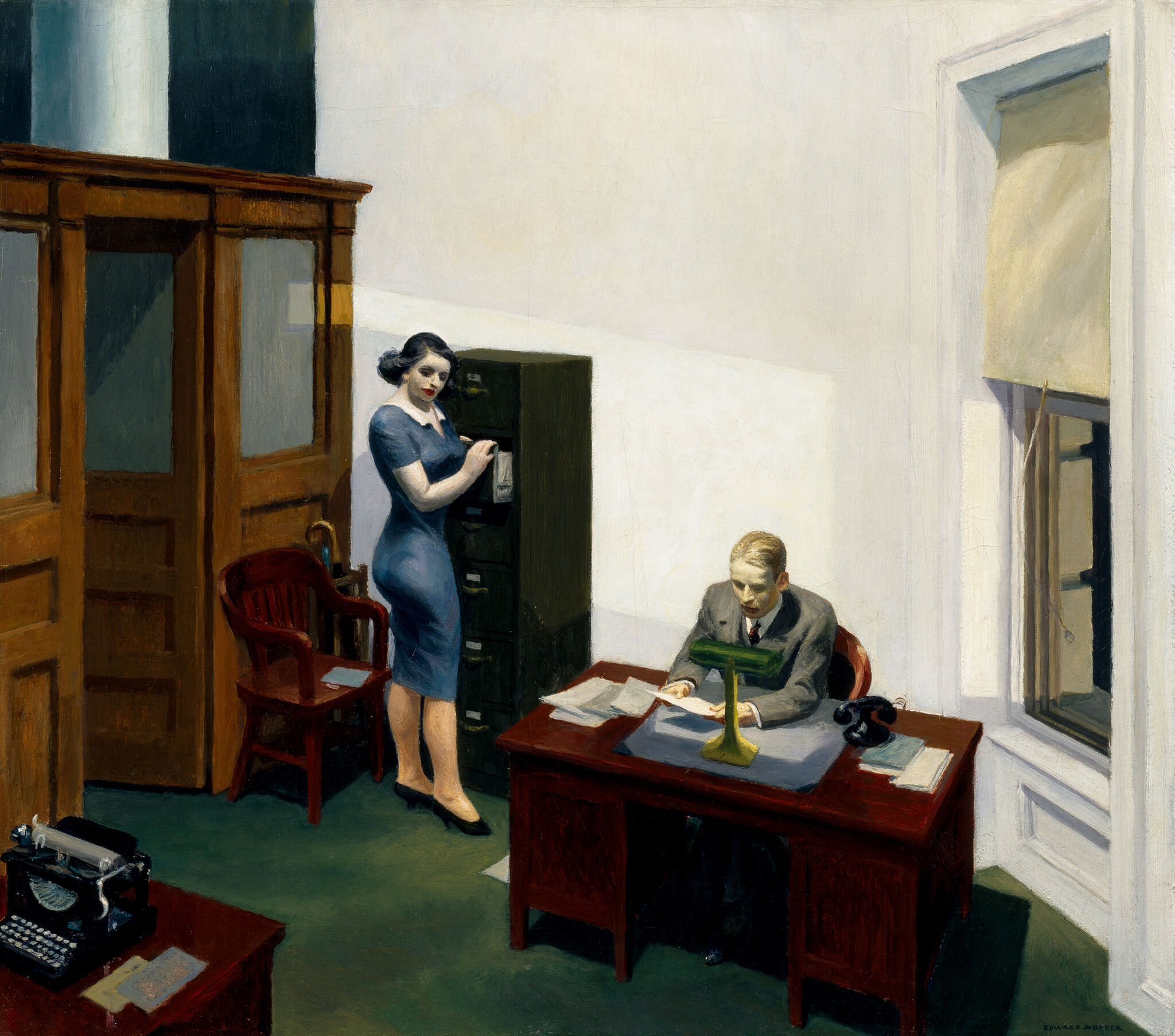 In an office a man sits at a desk looking at a piece of paper with a woman standing next to him. 