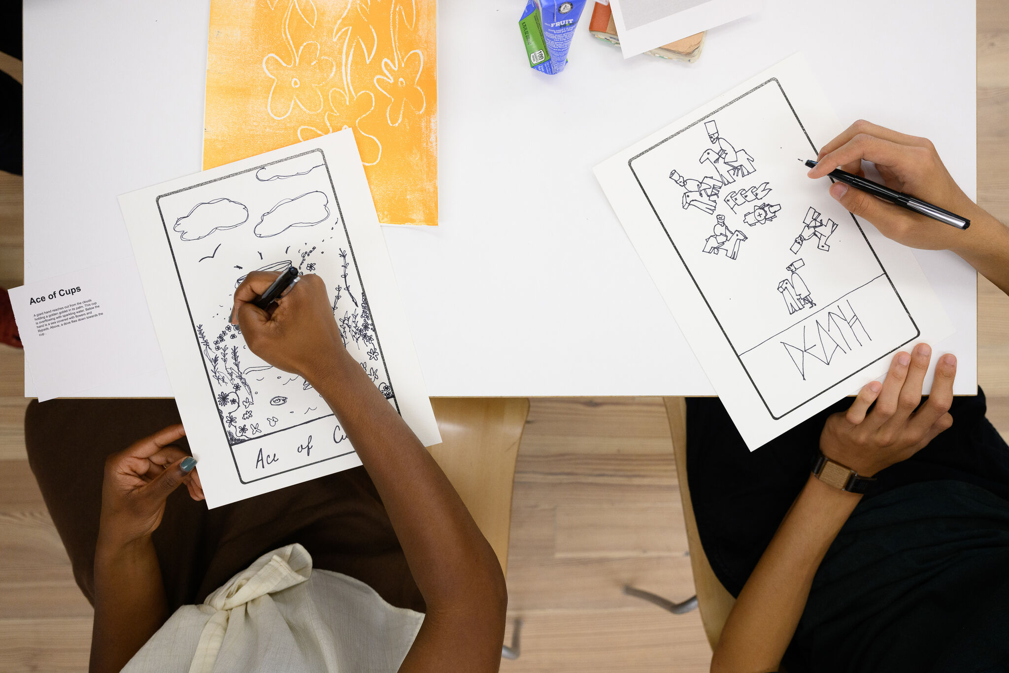 Students sit side by side.  They each have one sheet of paper and are using black markers to draw images.  In the background, there are colored pages and art materials on the table.