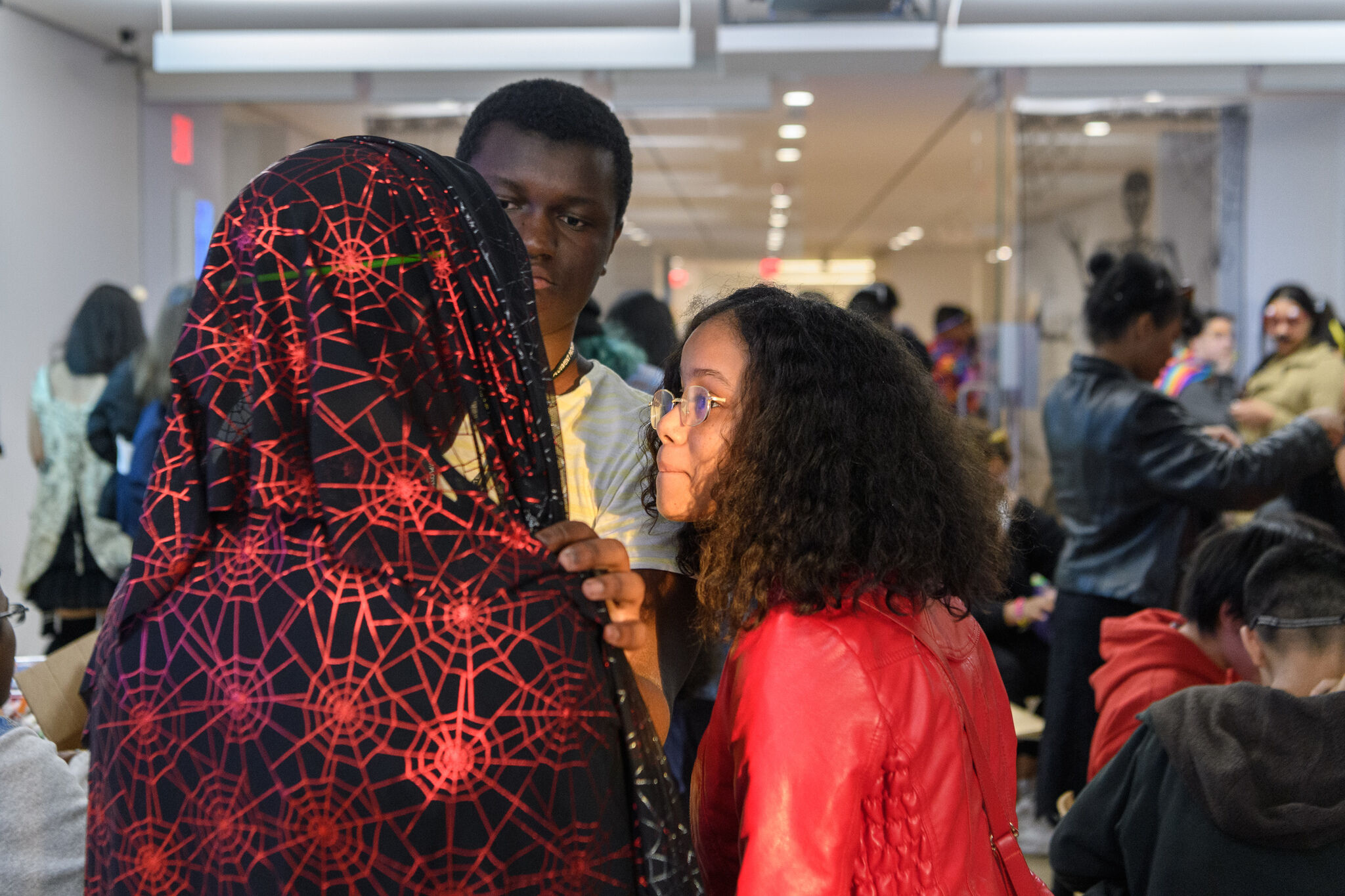 Teens create their own costumes in a workshop.  One person drapes a spider web pattered fabric over the other person, while an onlooker smiles.  In the background, teens are busy cutting and making costumes.