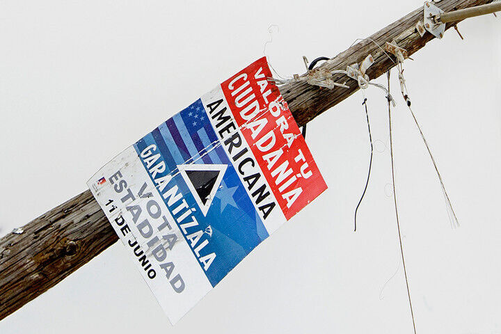 A worn red, white, and blue sign with text and images of Puerto Rican and American flags attached to a wooden post with wires attached to it.