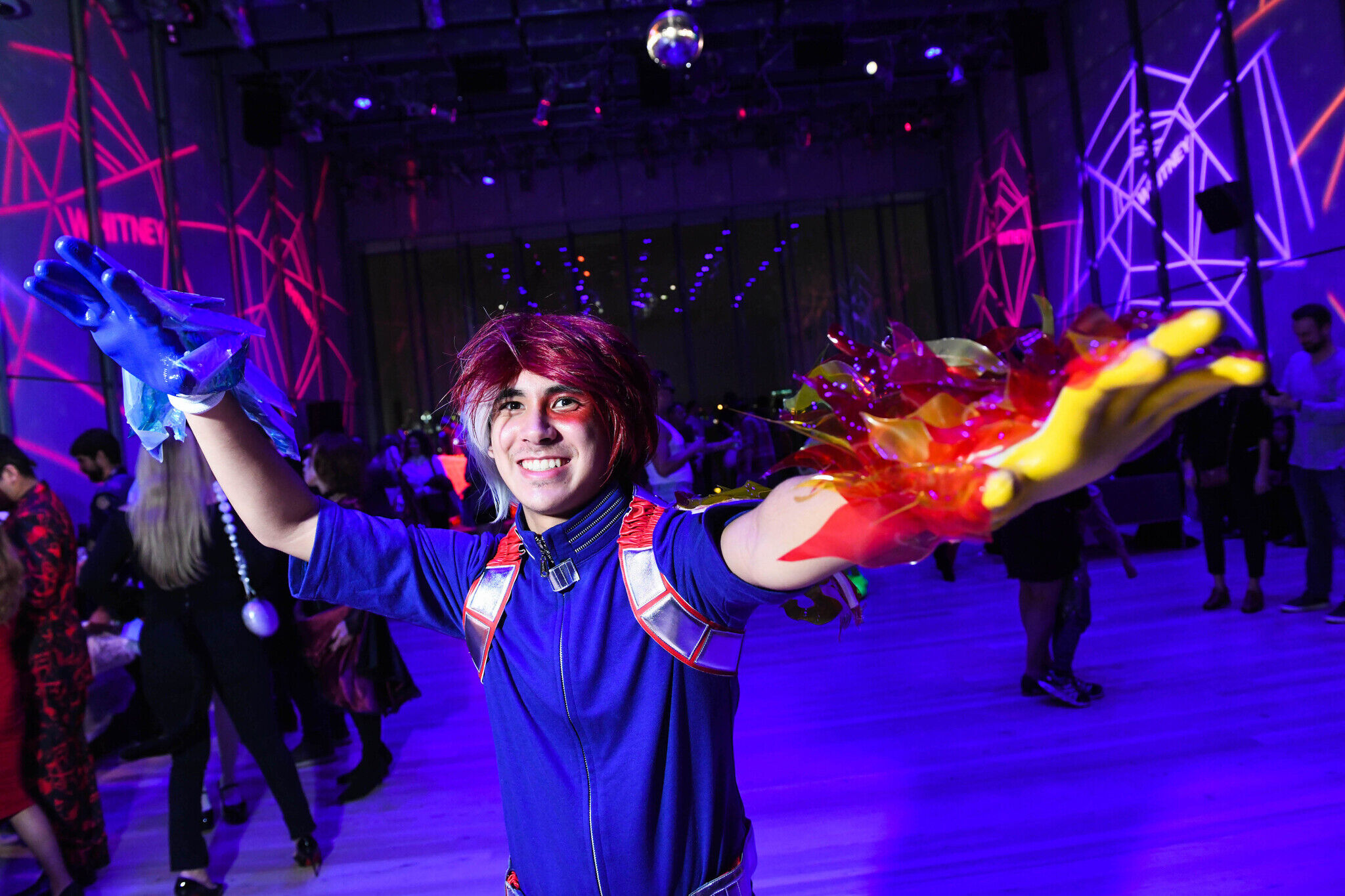 Person dressed up in costume with a right glove depicting water and left glove depicting fire. There is a disco ball on the ceiling and spiderweb shapes projected onto the walls of the room. There is a group of people in the background.