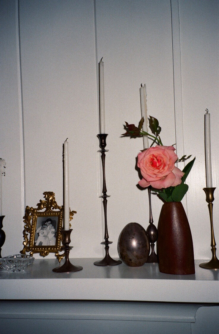 A photograph of a shelf supporting candles, candlesticks, a vase and rose, and a framed photograph. 