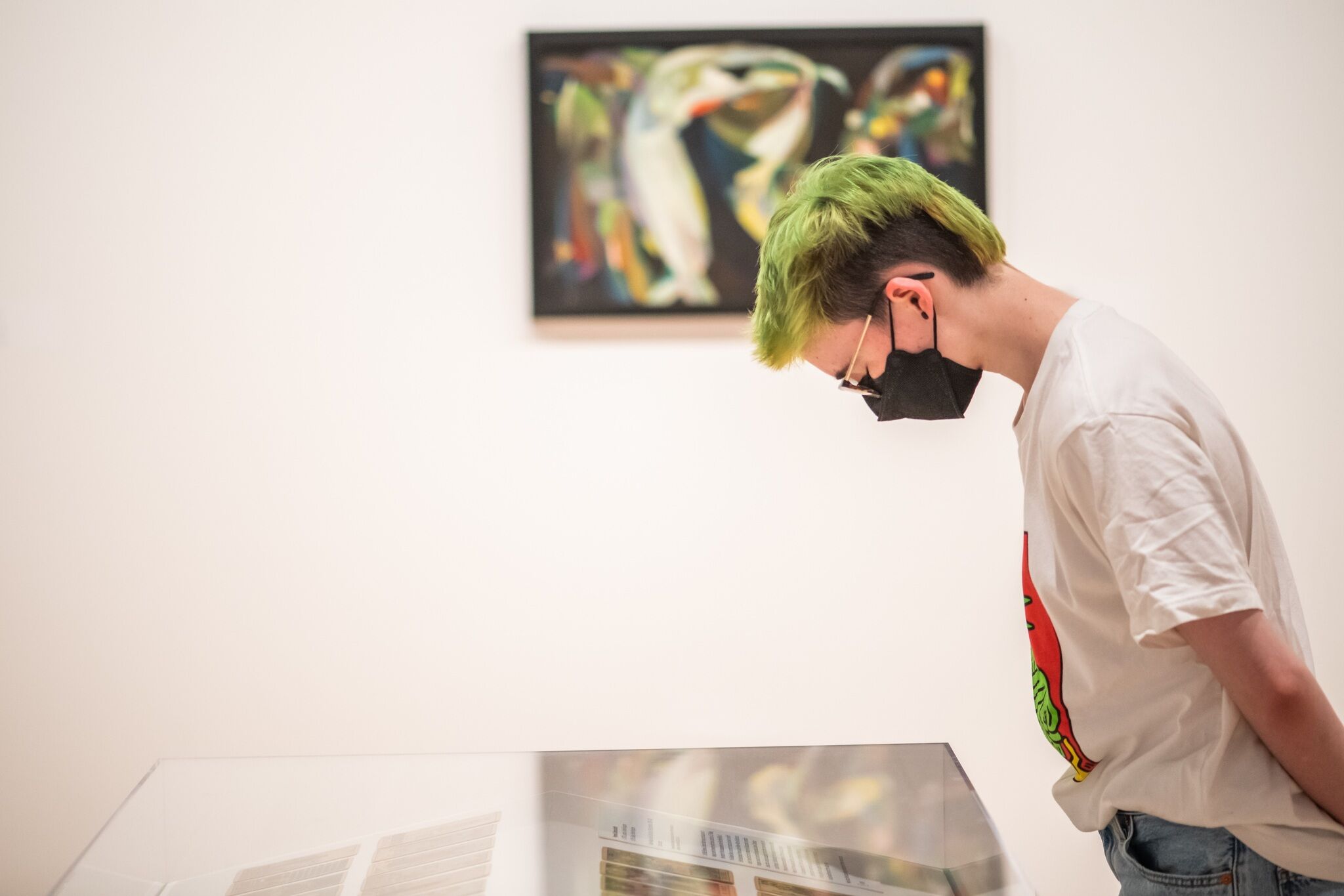 A person wearing a white shirt, black facemask, and glasses looks down at a glass case with small cards inside in a gallery with white walls. There is an out-of-focus abstract artwork hanging on the wall behind the person.
