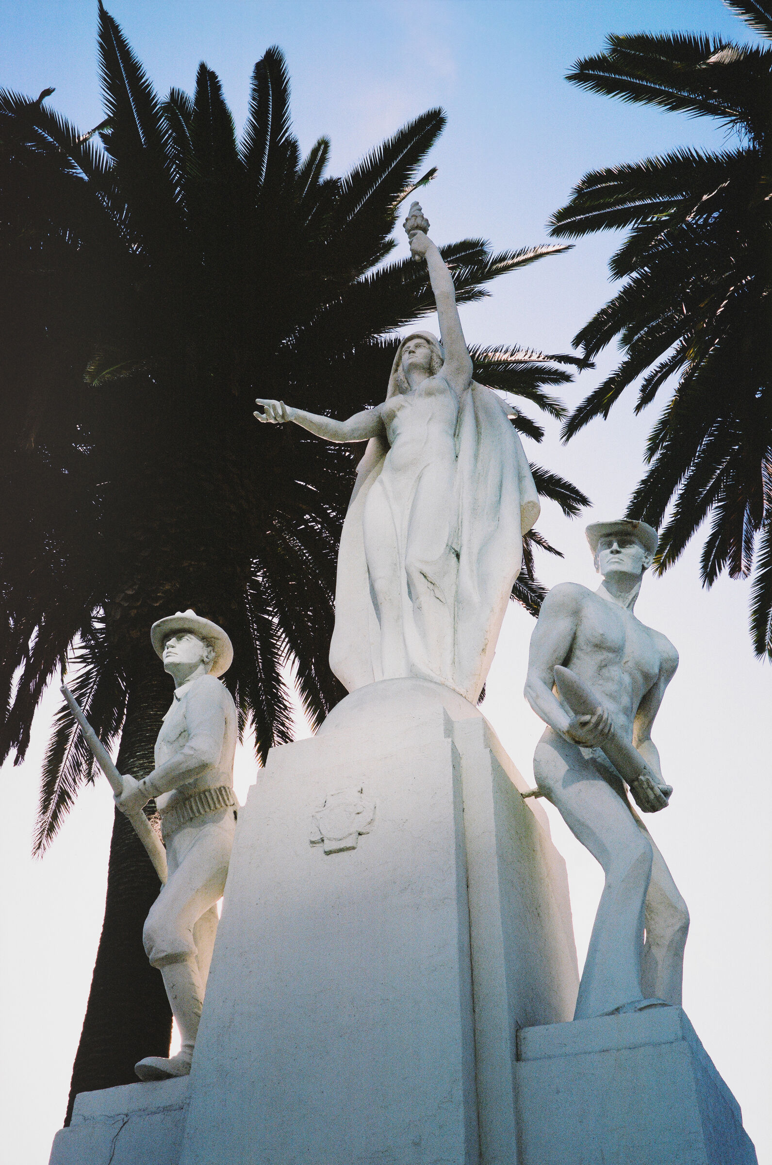 A photograph of three white sculptures of people taken from below with palm trees in the background.