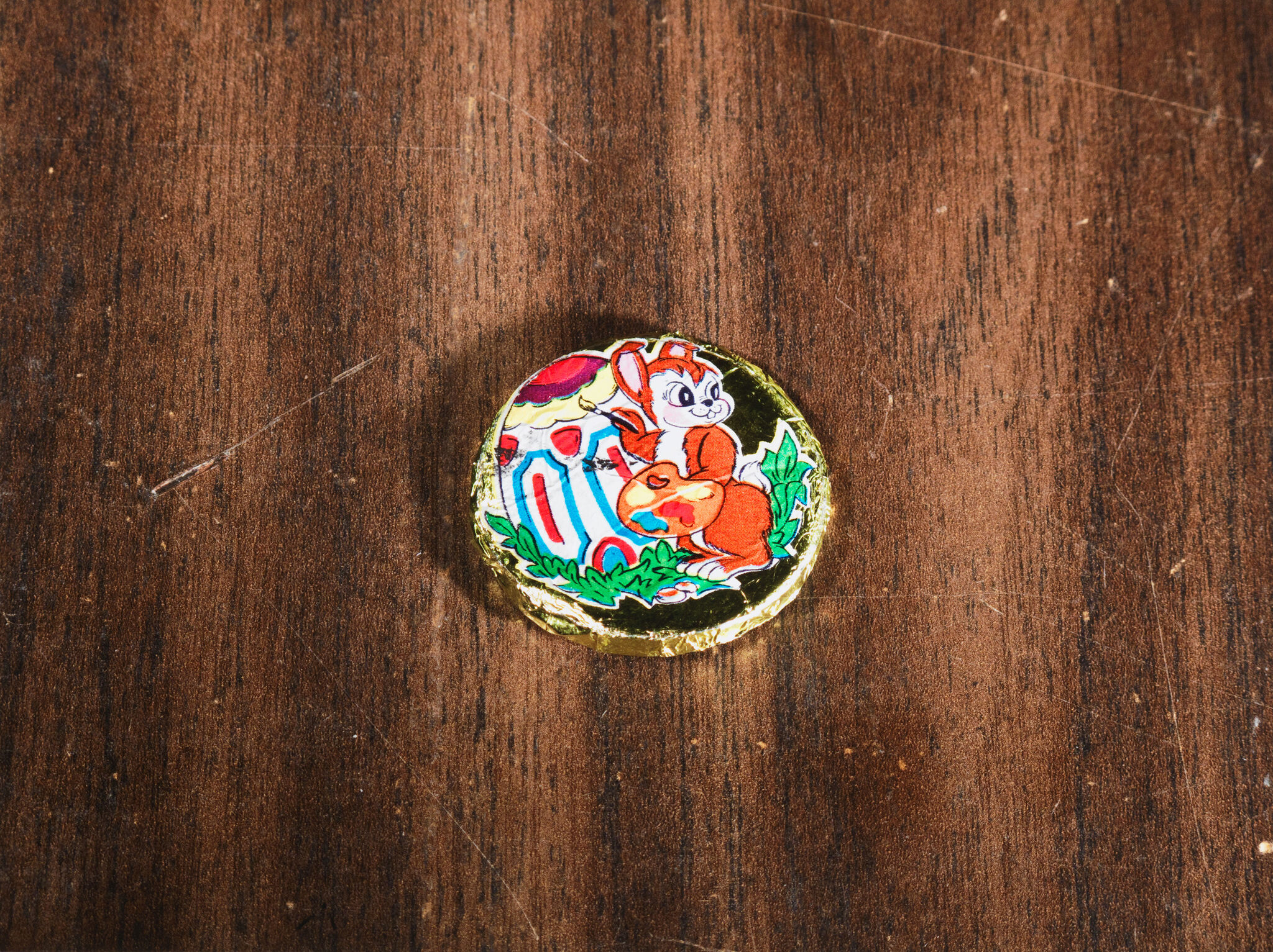 A photograph of something circular and flat lying on a wood table. The item is covered in gold foil printed with a colorful cartoon of a rabbit painting.