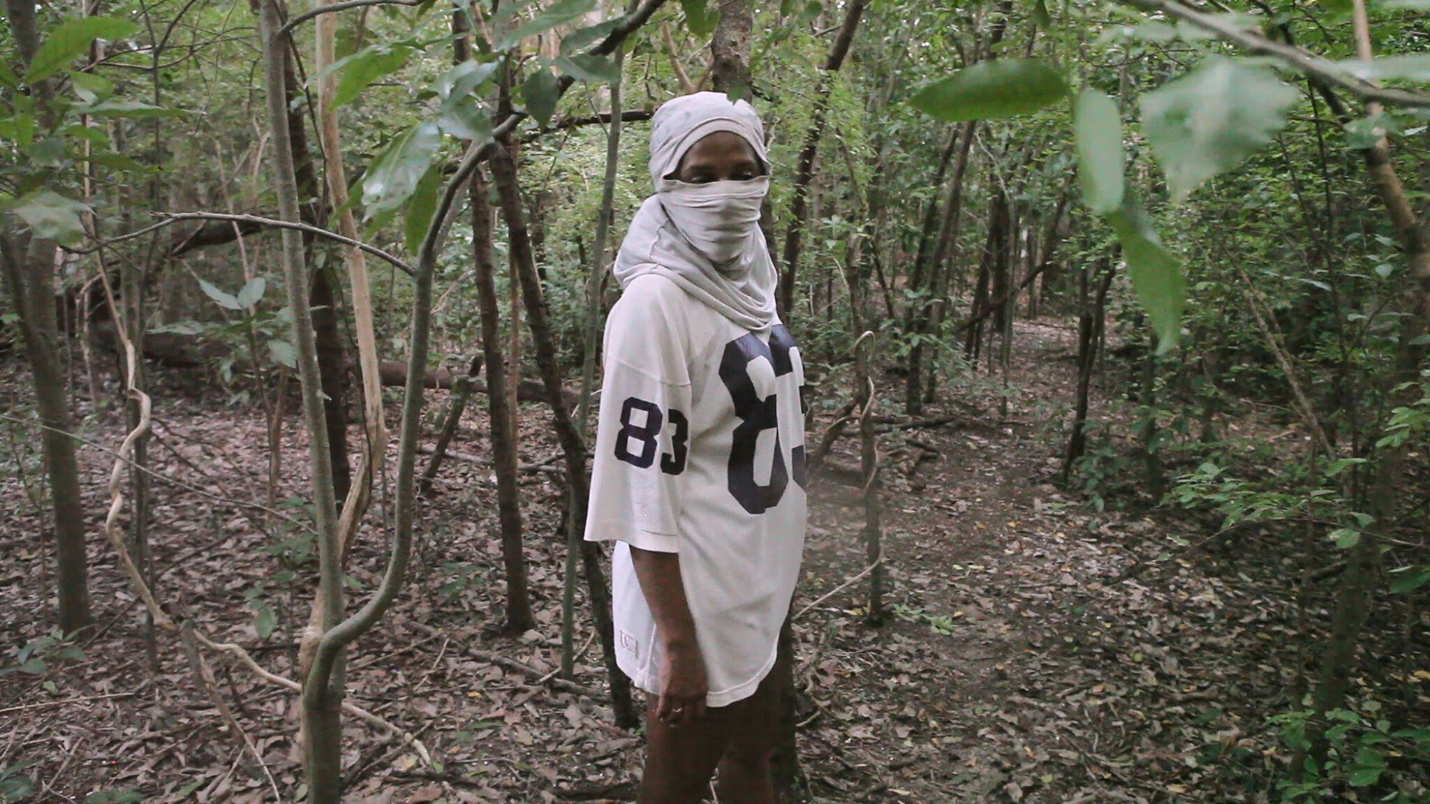 A many in a wooded area faces the camera with his face covered in cloth. 