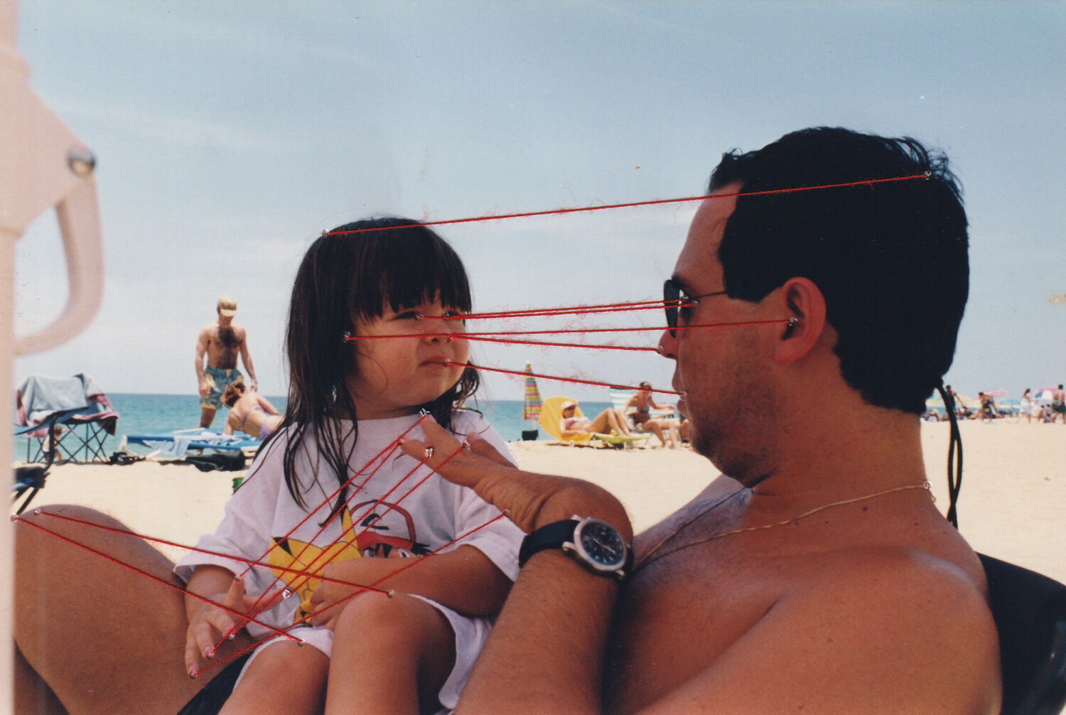 A photograph of a young girl sitting on the lap of a man at the beach. Lines of red thread are attached to the photograph.