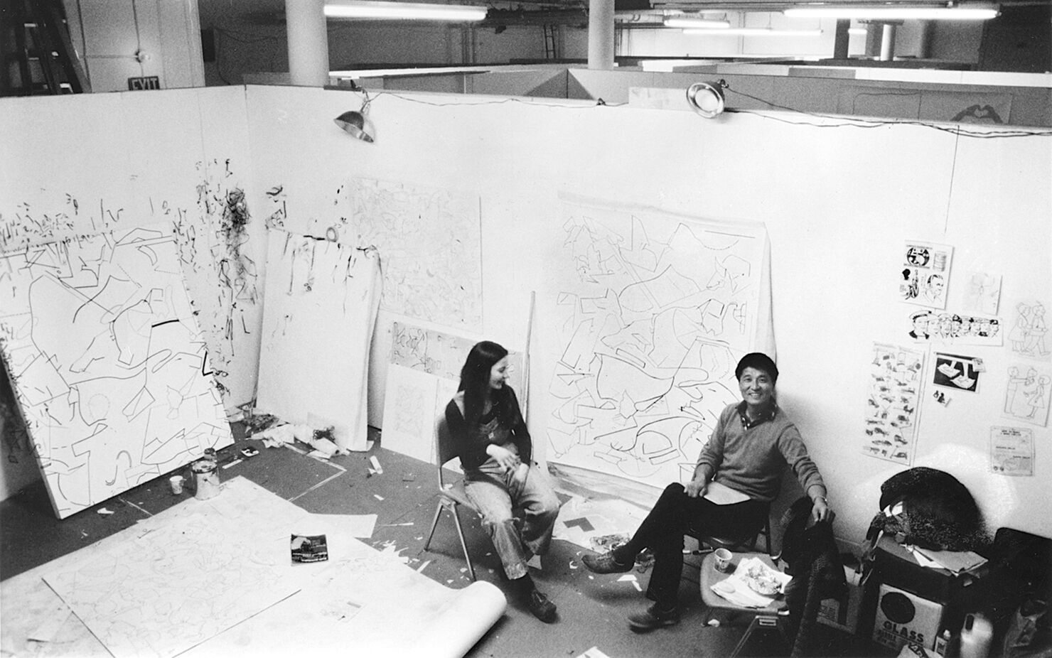 Two people sitting in an artist studio surrounded by paintings.