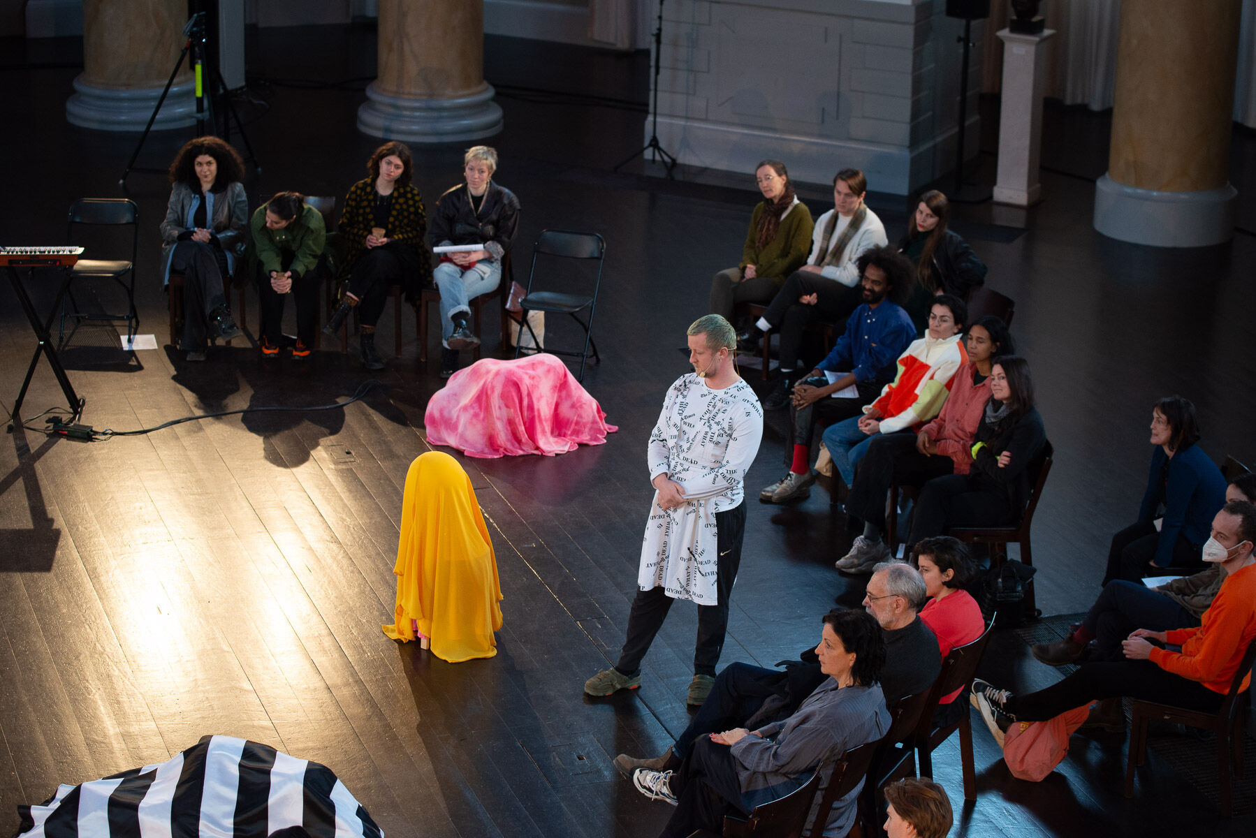A circle of audience members surrounds three performers. One is shrouded in a gold-colored fabric, and another in a pink tie-dyed fabric, with both crouched on the black wooden floor. The third performer, with blue hair and a patterned black-and-white top, stands and looks to their right. 