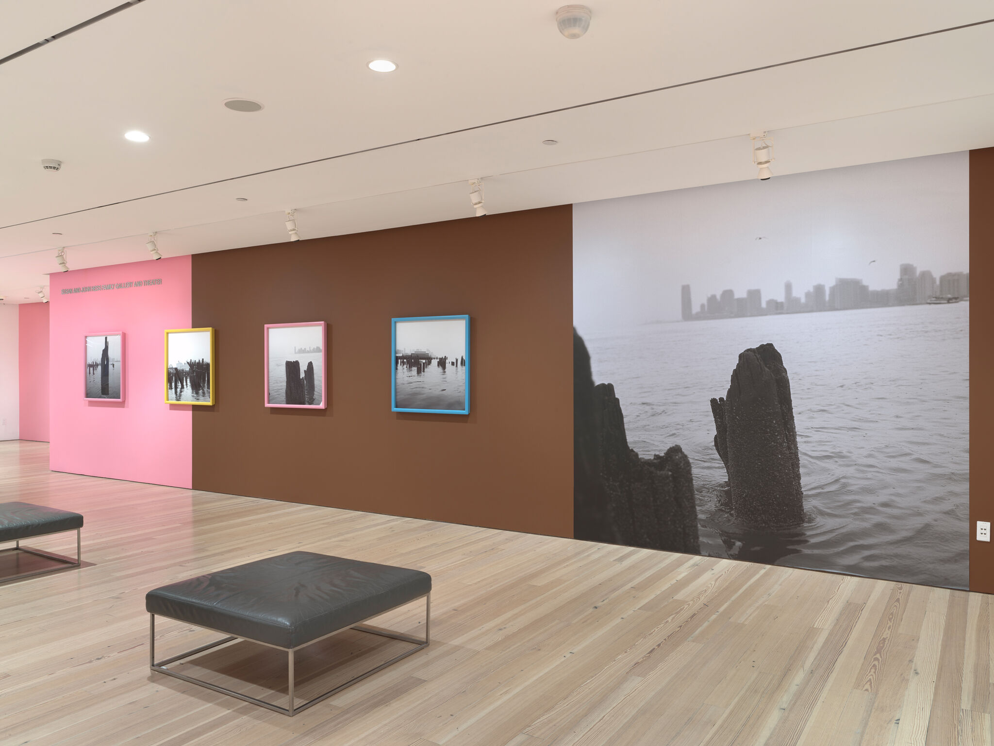 Four black and white photographs of Manhattan's westside waterfront on a pink and black gallery wall next to a large photograph of the Hudson River