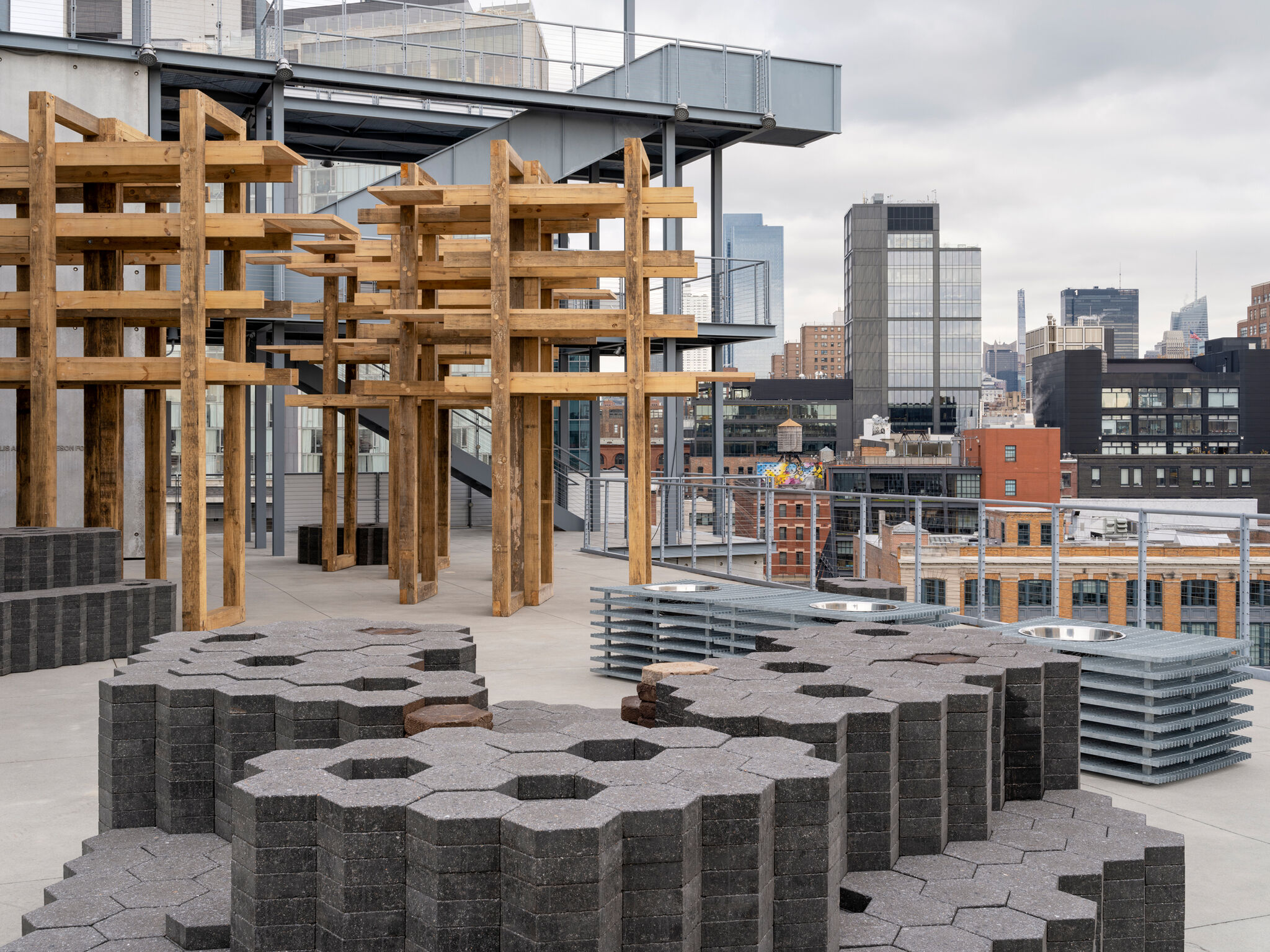 Several stacks of hexagonal, coffee-clay pavers and tall wooden-beamed structures installed outdoors on the Whitney's terrace, with a view of the New York skyline behind. 