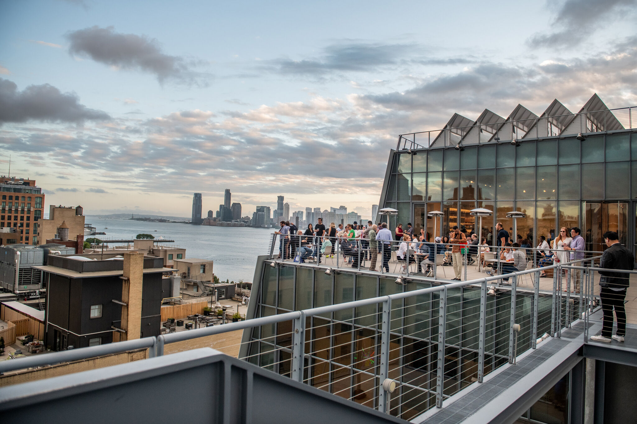 Exterior view of the Whitney Museum terrace at dusk with pink, blue, and gray clouds above the Hudson River and New Jersey skyline. Dozens of members mingle on the eighth floor terrace overlooking NYC.