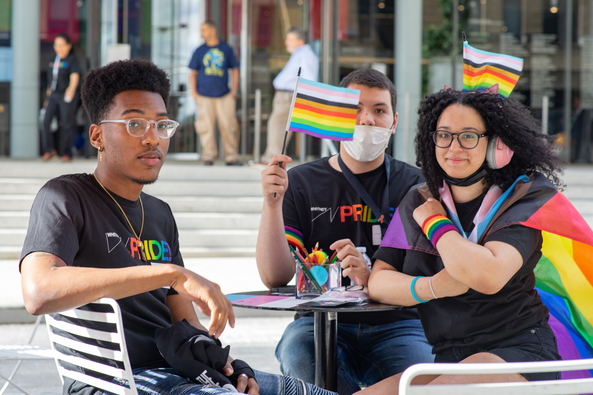 Three youth smile at the camera while wearing inclusive pride flags.