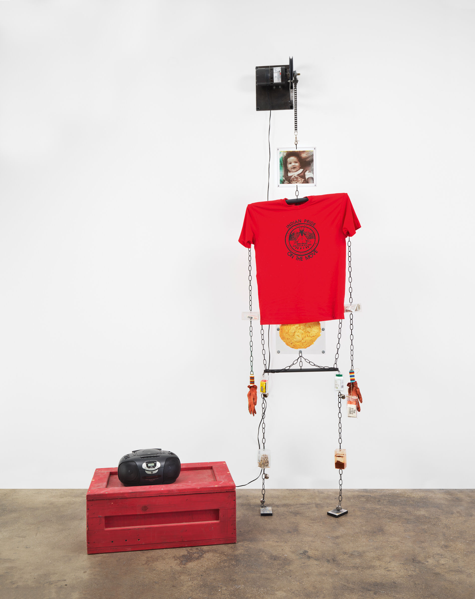 The shape of a human constructed out of chains, gloves, a t-shirt, and a photograph of a child's face next to a boombox. 