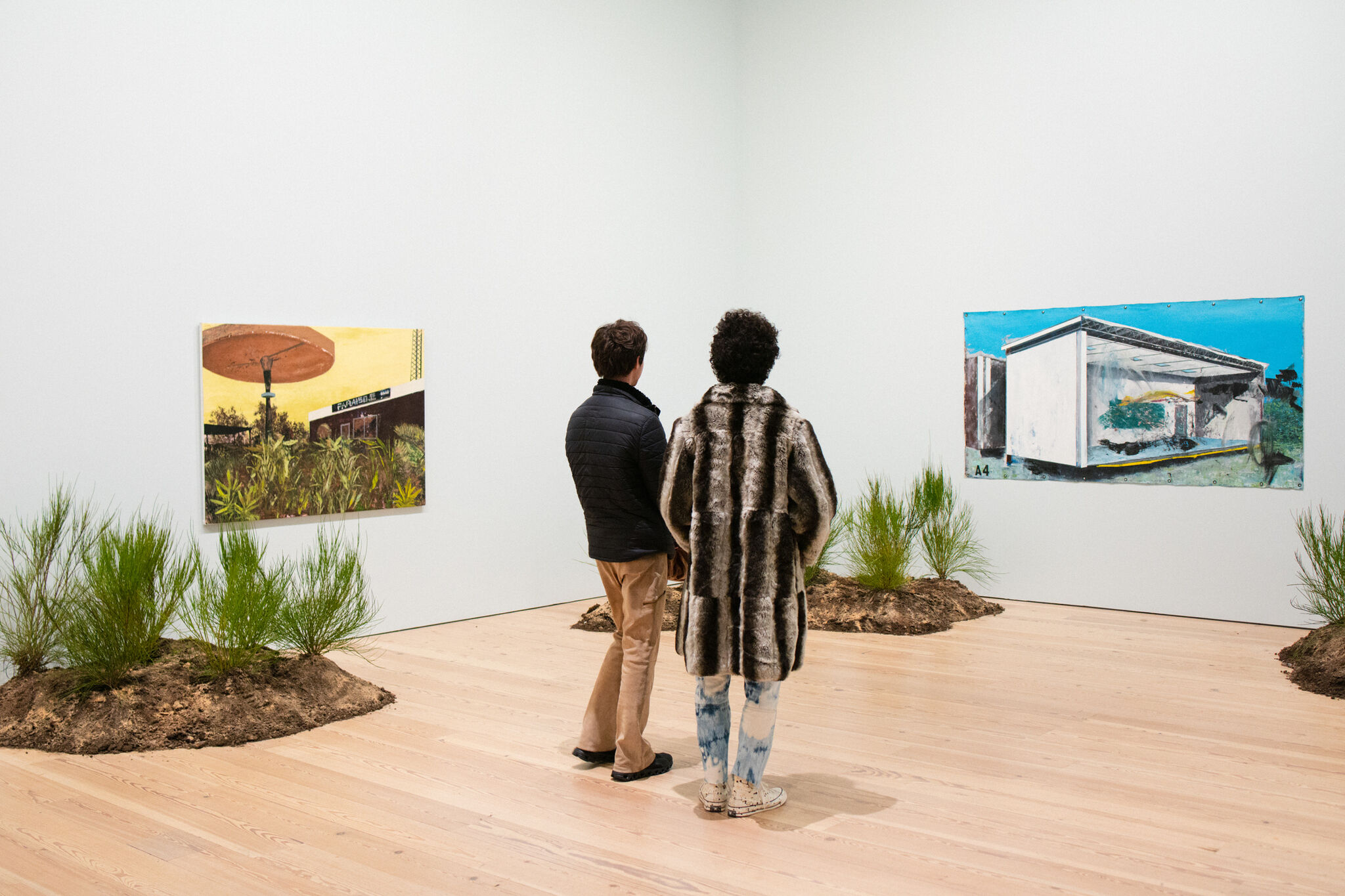 Two Whitney Museum guests–one wears a black jacket, the other wears a striped fur coat –stand in the middle of a gallery facing an installation by artist Rogelio Báez Vega depicting abandoned buildings in Puerto Rico. On the gallery’s wooden floors, there are small green shrubs growing out of mounds of dirt.