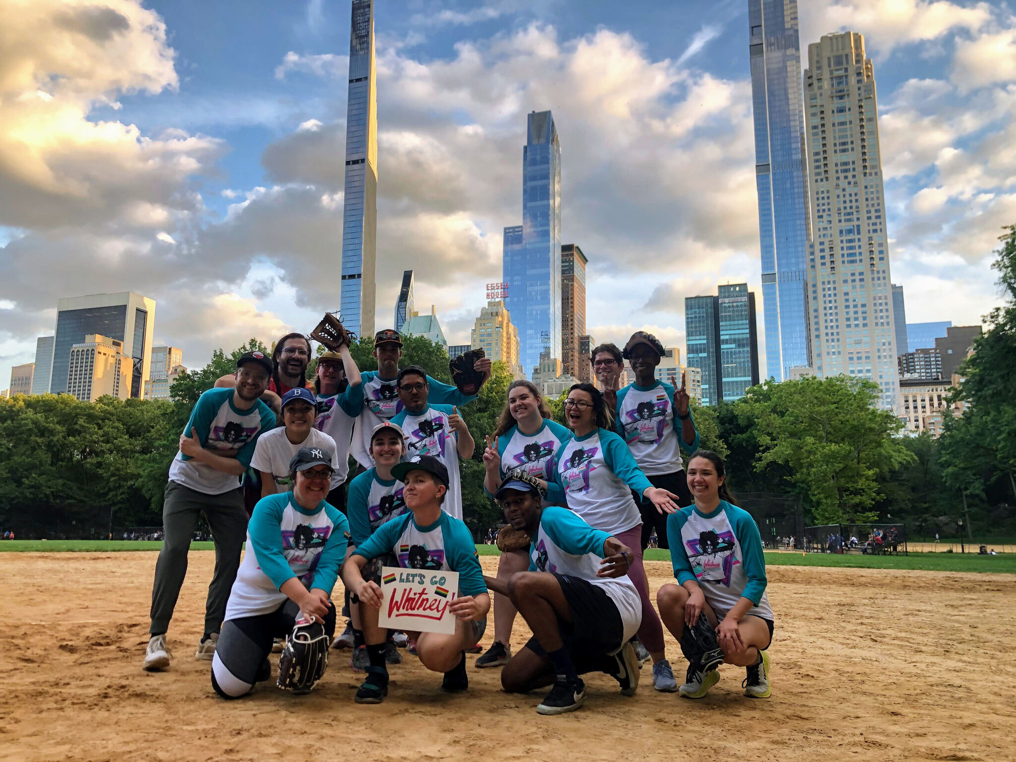 A group of people stand around a sign that reads "let's go Whitney" in a softball field with the New York City skyline behind.