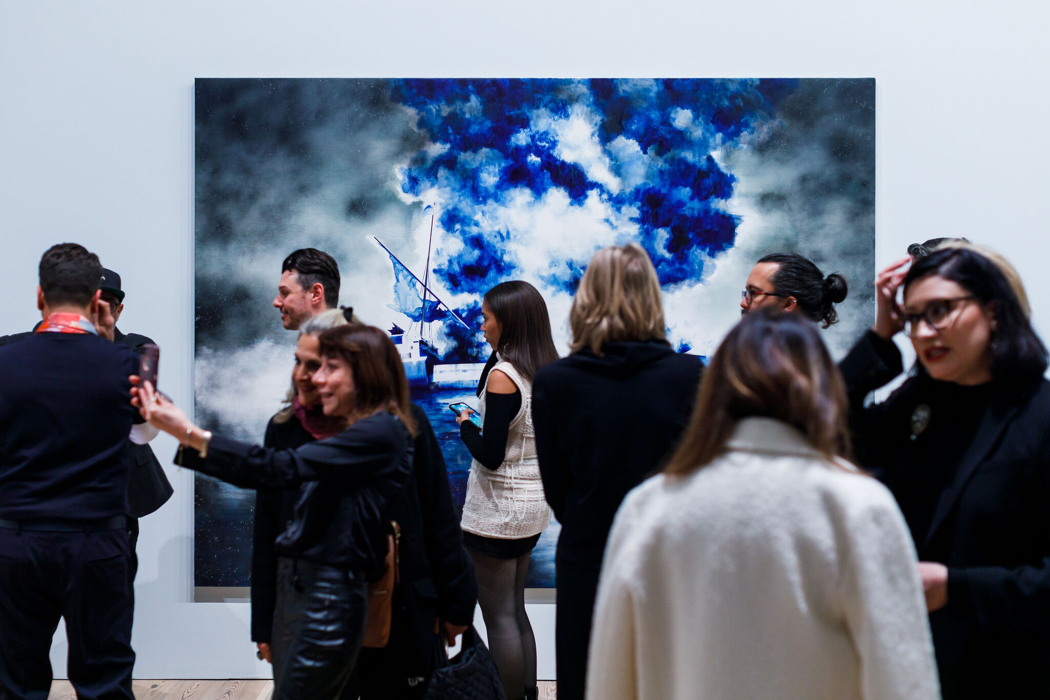 A group of people scattered in the gallery looking at artwork and talking with one another. In the background is a largescale artwork featuring a ship, sky and clouds