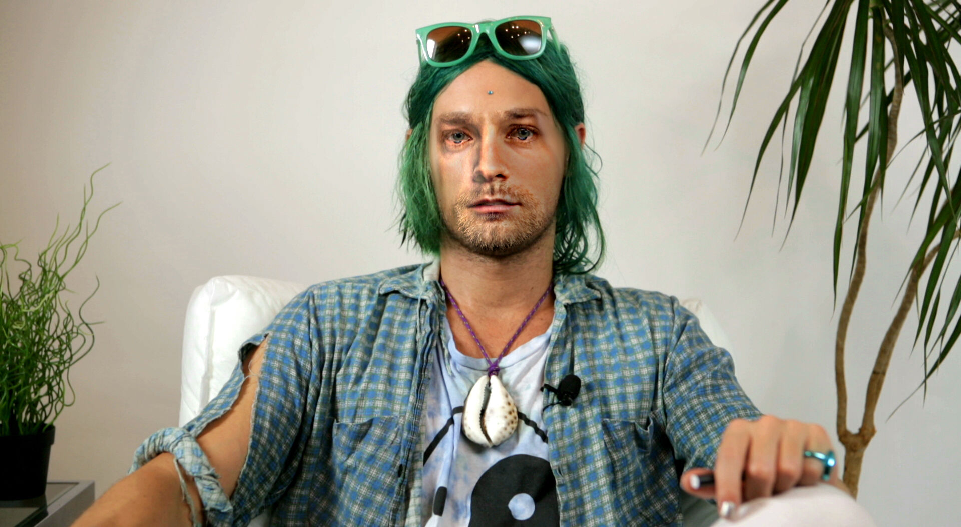 A man that looks like Kurt Cobain sits on a chair looking at the camera. 