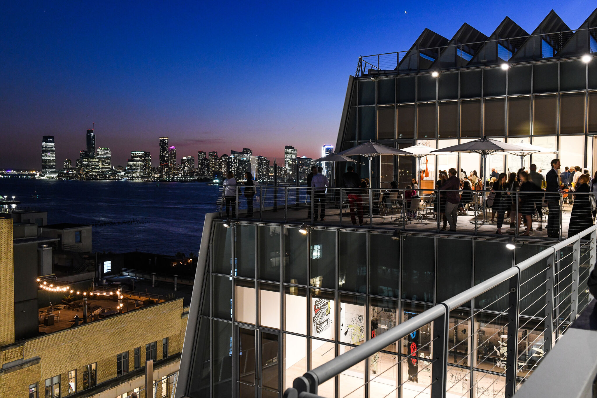 Outdoor terraces and staircases at the Whitney Museum. People walk around and point to the view of the city around them. Through the windows are the art galleries, and the cityscape reflects in the windows.