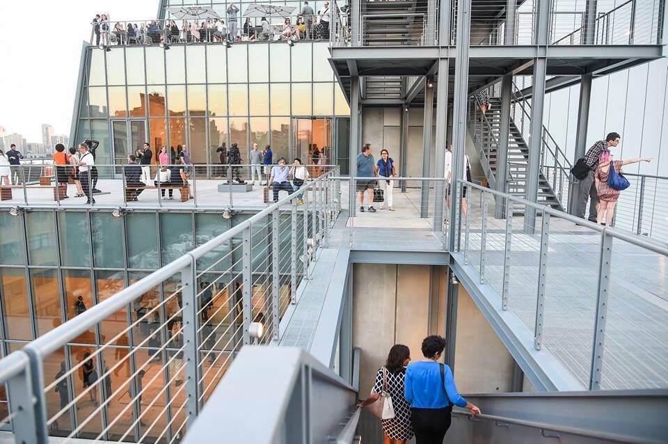 Outdoor terraces and staircases at the Whitney Museum. People walk around and point to the view of the city around them. Through the windows are the art galleries, and the cityscape reflects in the windows.