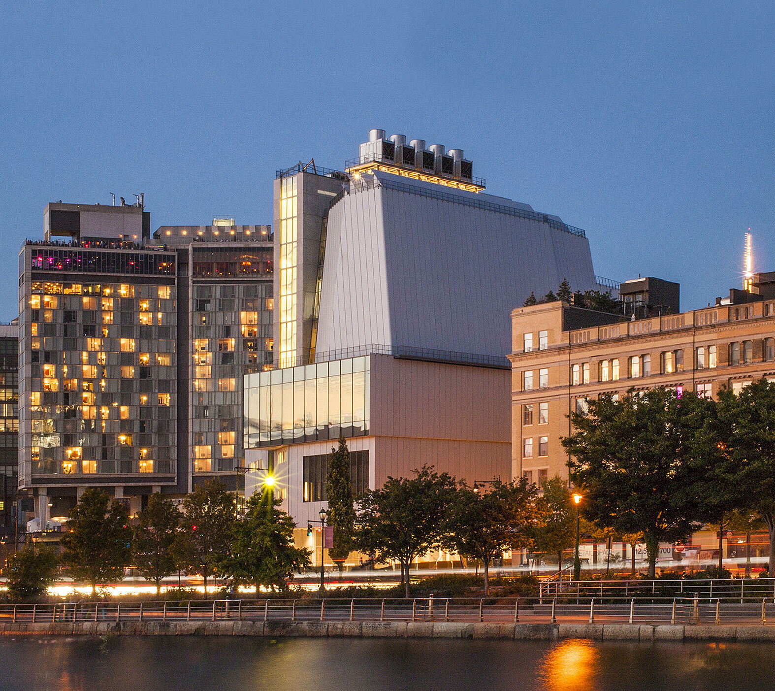 Whitney Museum of American Art and surrounding buildings with sunset light hitting their facades. The river is visible in front of the buildings, and the sky is bright.