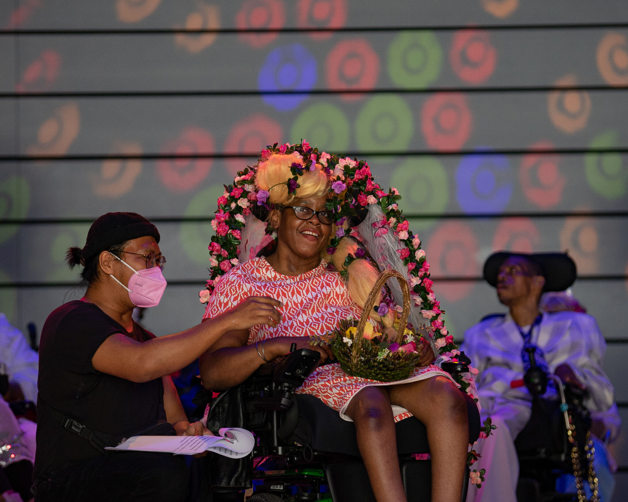 A Black person in a wheelchair and eyeglasses wearing a large flower crown smiles widely. 