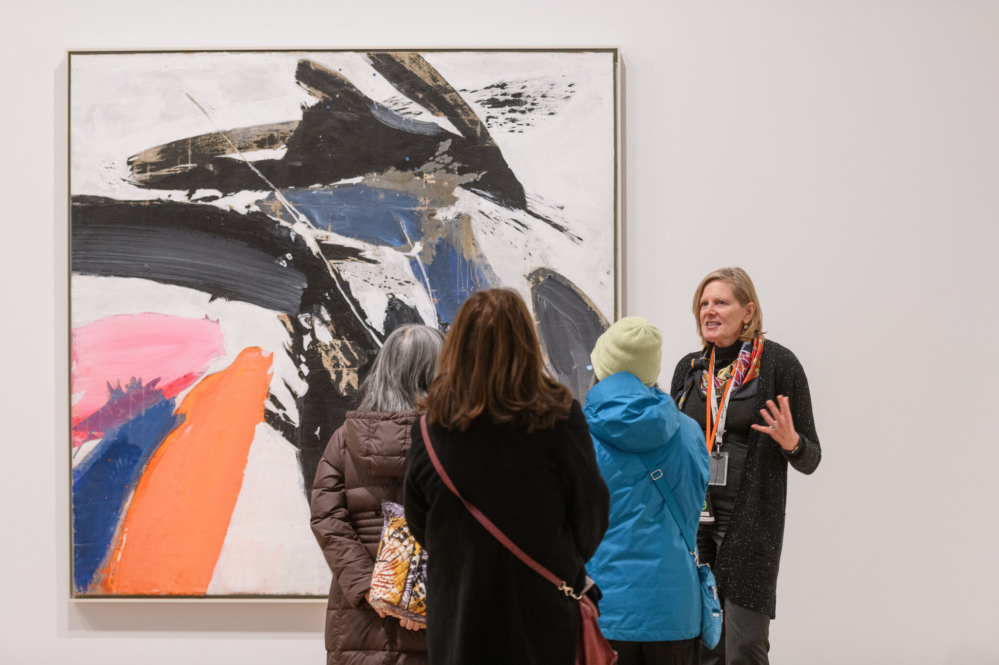 Three people stand around a large painting with swaths of black, orange, pink, and blue.