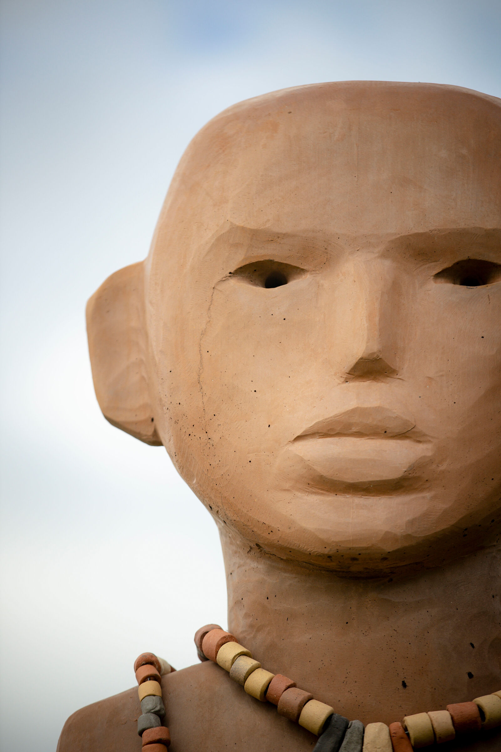 A figurative sculpture stands against the sky with holes for eyes and a clay surface. 