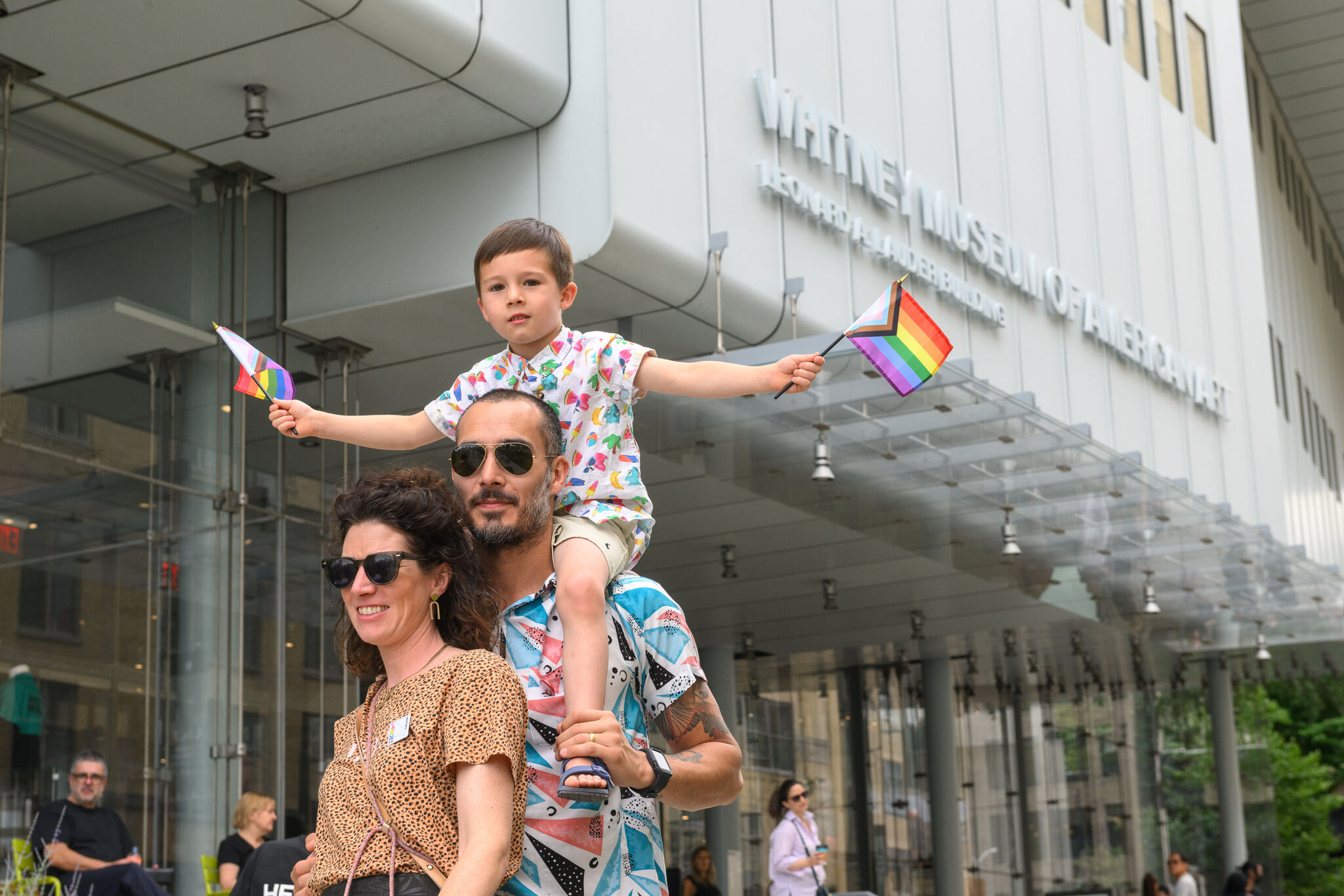 Two parents in front of the Whitney's entrance. One of them has a child holding Pride flags on their shoulders.