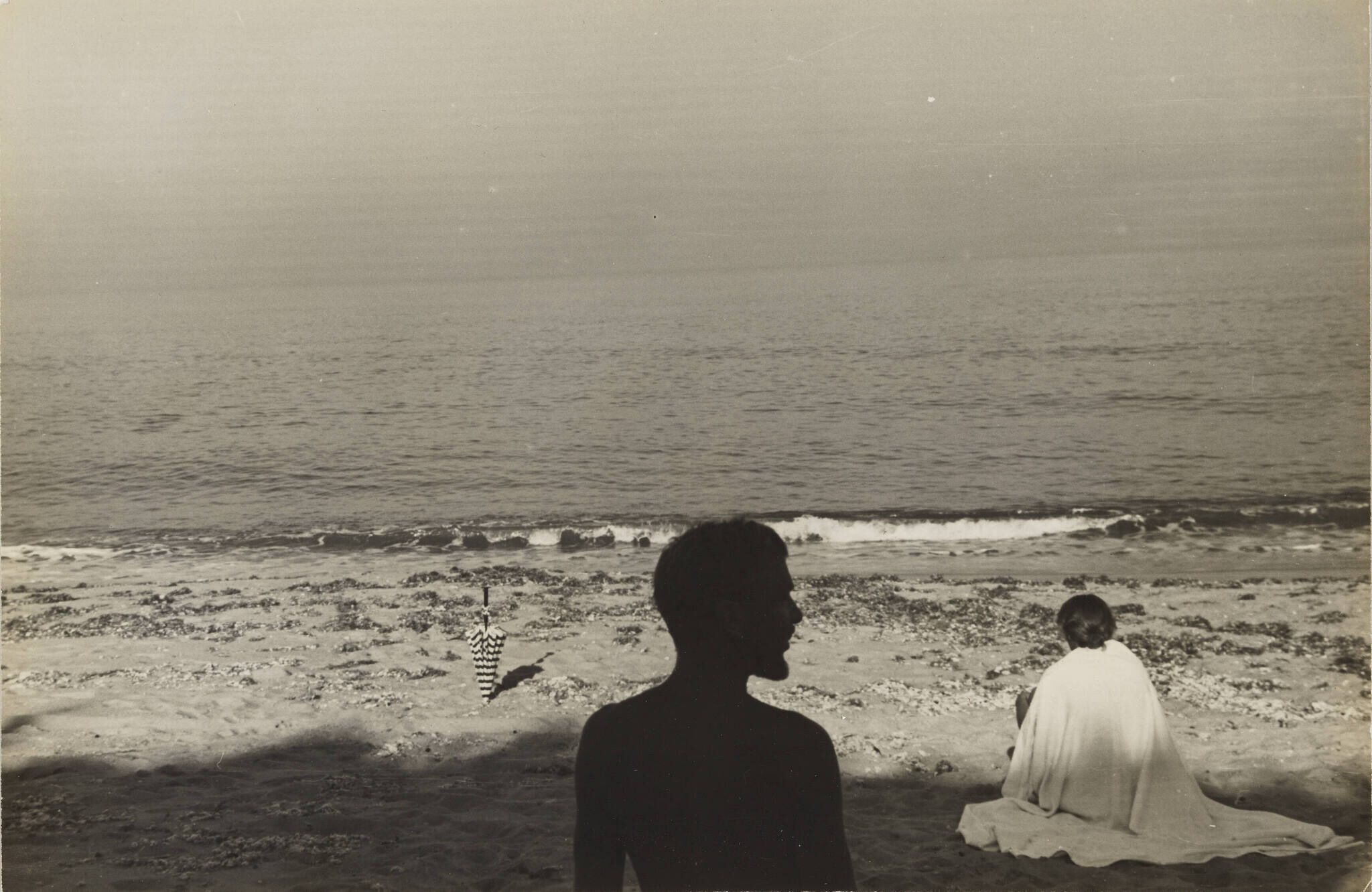 A black and white photograph of two people on the beach. The man is in the center of the frame in total shadow and silhouette, while the woman sits on the sand in a flowy white garment with her back to the viewer. 