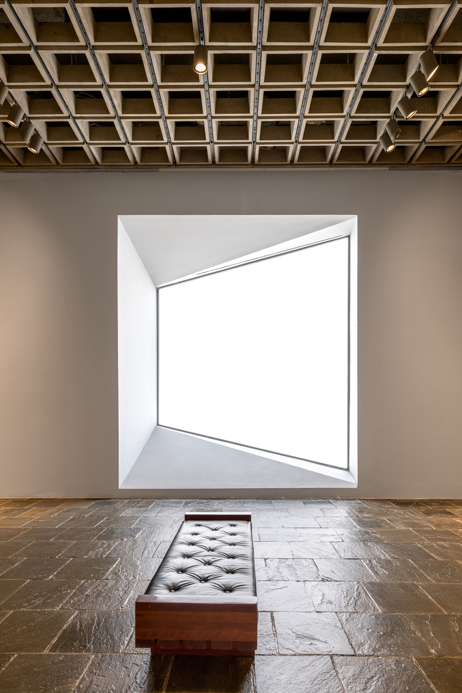 Interior gallery of the Breuer Building with light shining through a large window, a textured ceiling, and a leather bench.  