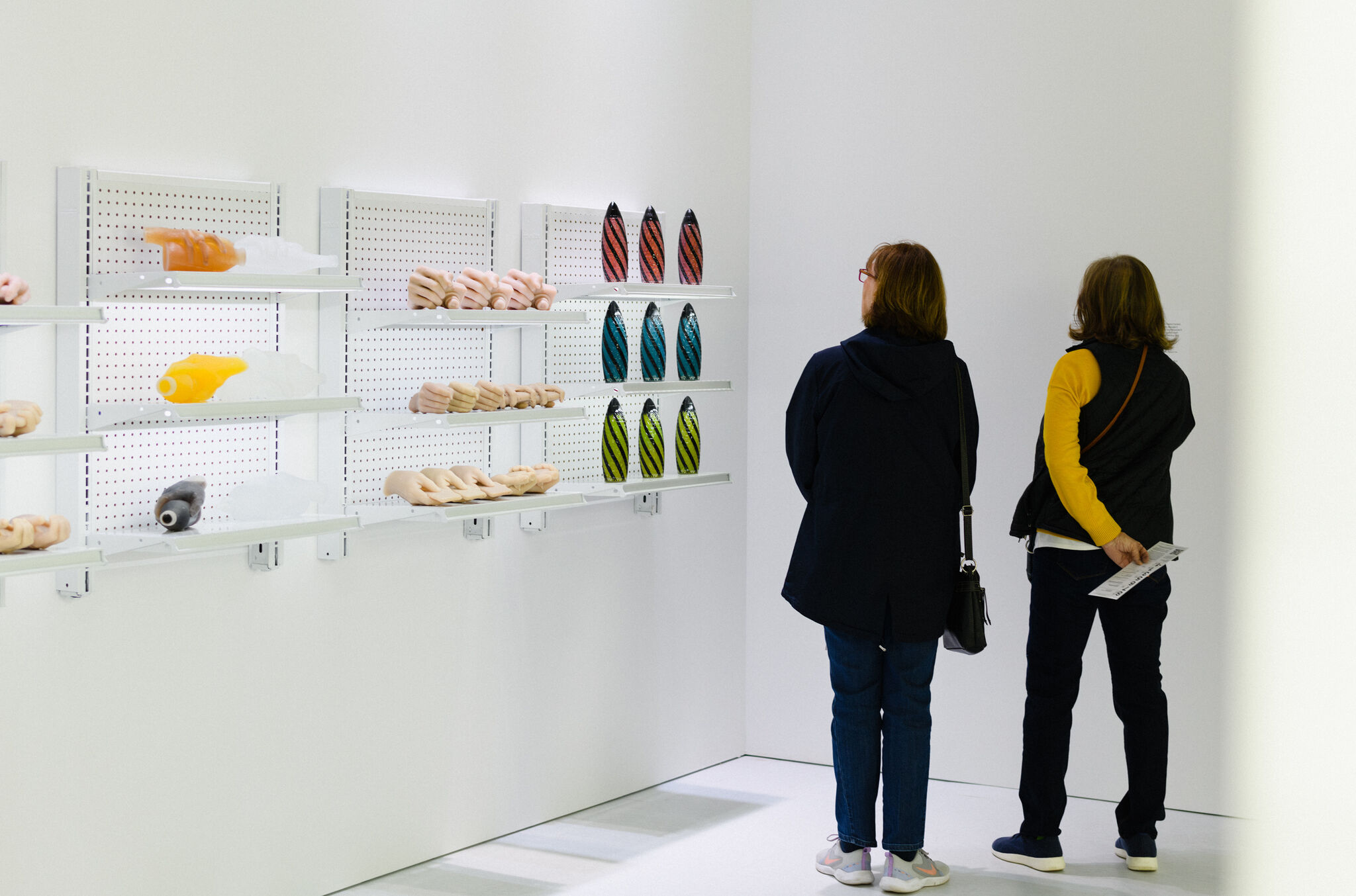 Two museum visitors stand in the corner of a gallery with industrial shelves on the wall filled with colorful sculptures, including multiple lifelike hands.