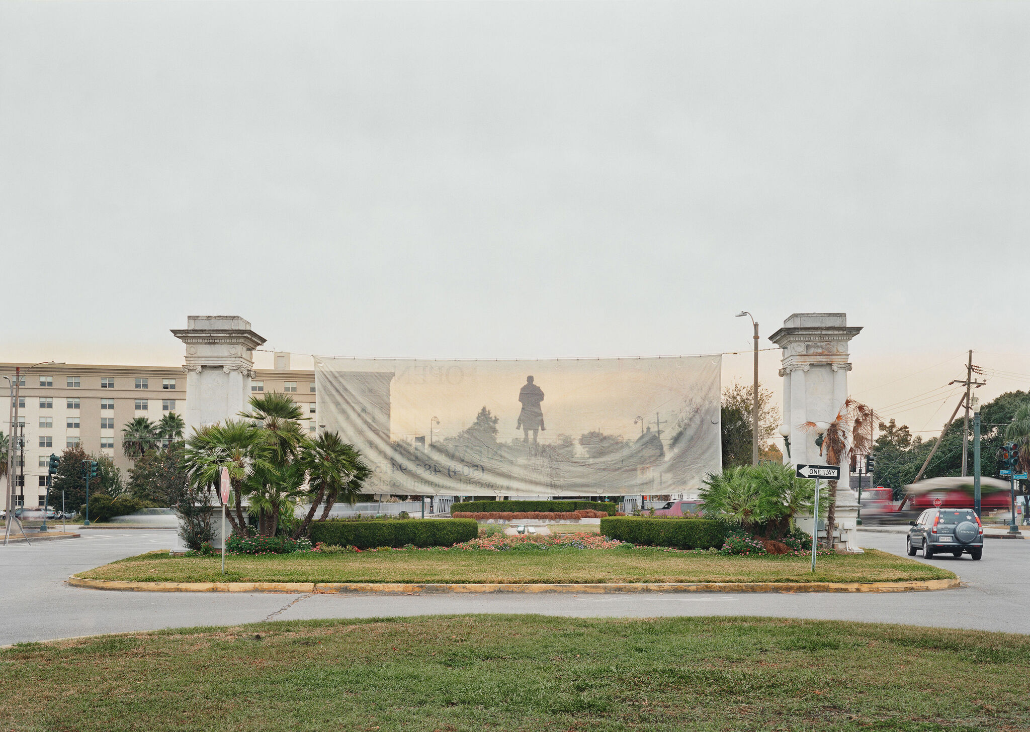 A monument of a man riding a horse behind a translucent banner hung between two columns on a grassy street island