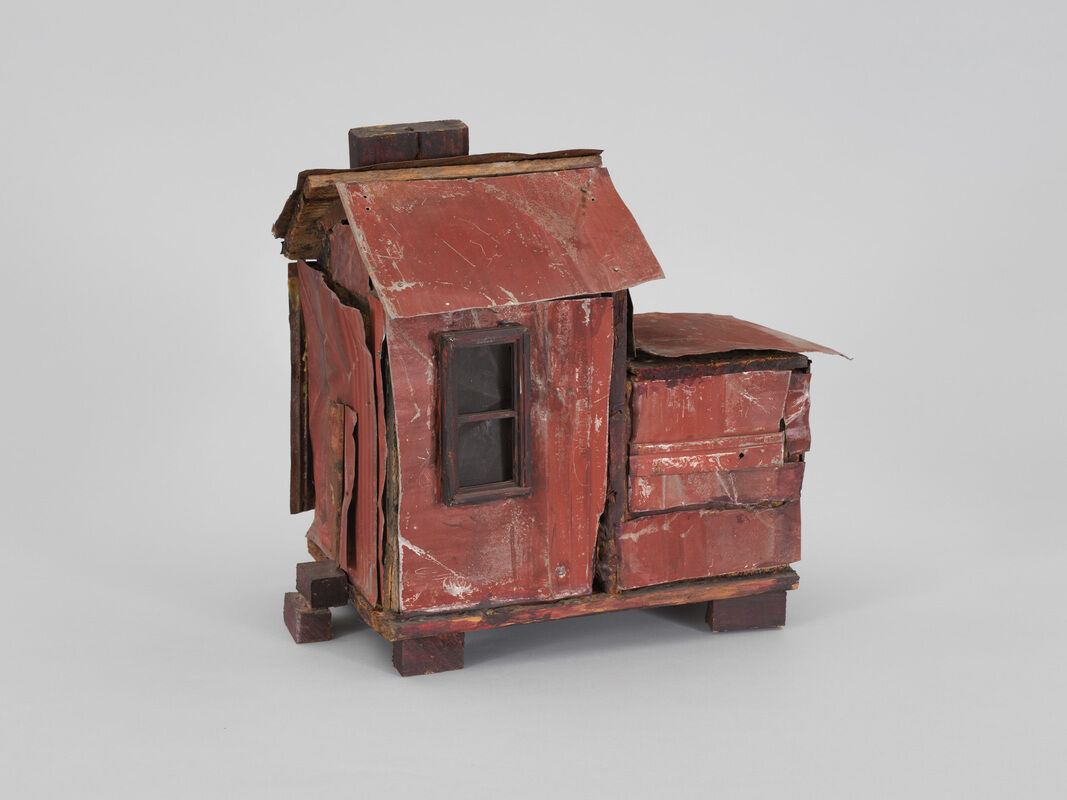 A rusty red and brown two foot tall house structure is sculpted from wood and tin. The exterior contains a small window, with opaque panes, and is dinged and scratched up. The house sits on two visible wooden planks which keep the foundation from sitting squarely on the surface.