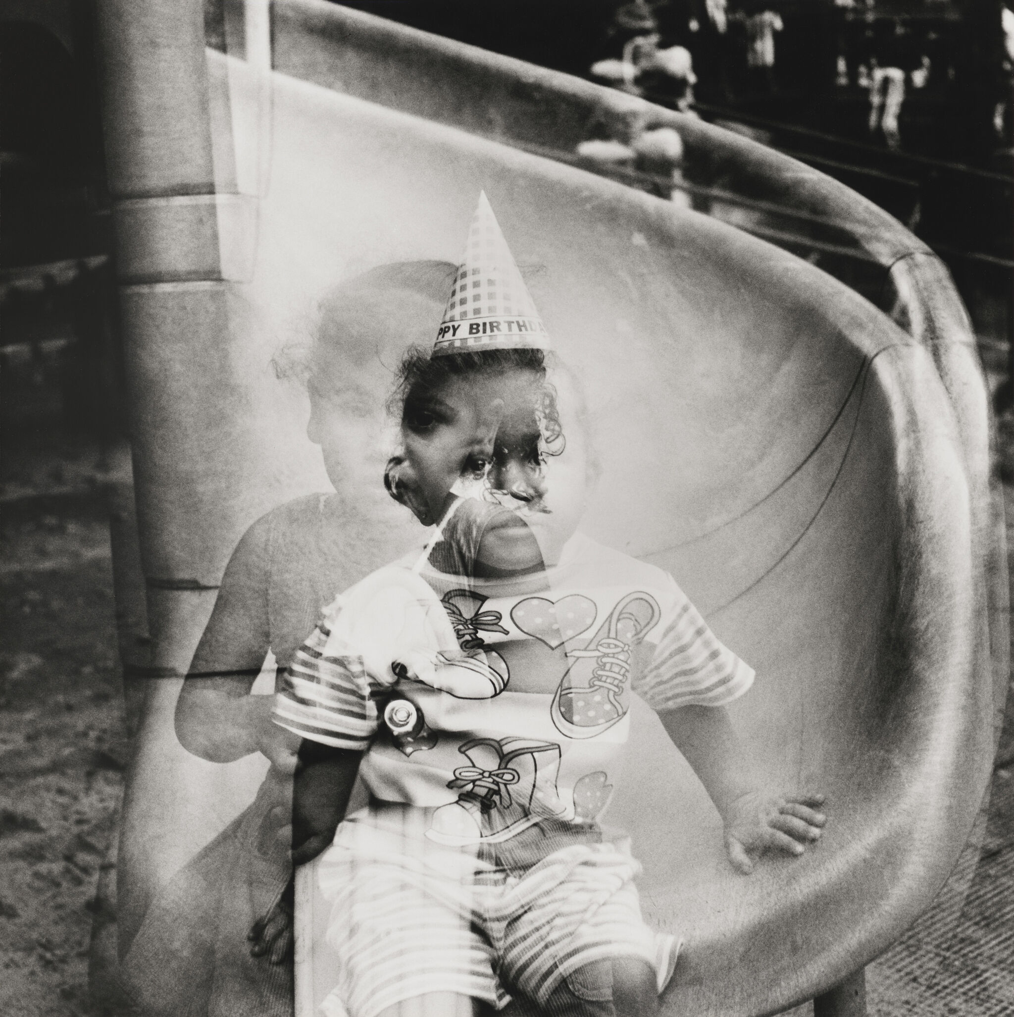 A black and white photo centers a baby sitting on the edge of a slide, wearing a conical birthday hat and holding theirself up by leaning on their right arm. Overlaid on top of the image of the baby is a ghostly, transparent image of a toddler, off-center to the left.