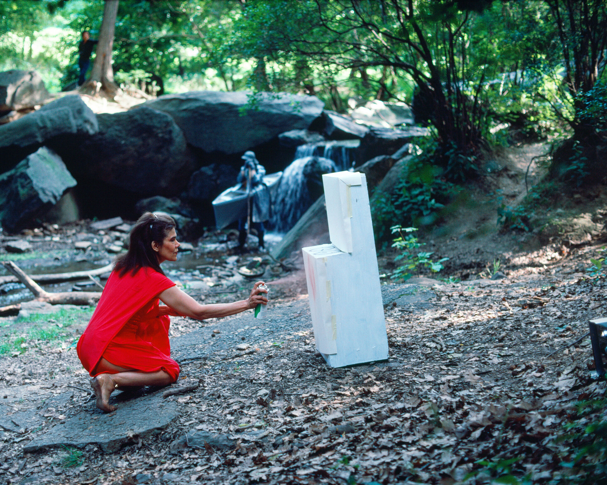 A woman in red bends down to spray paint a white object in the forest. 