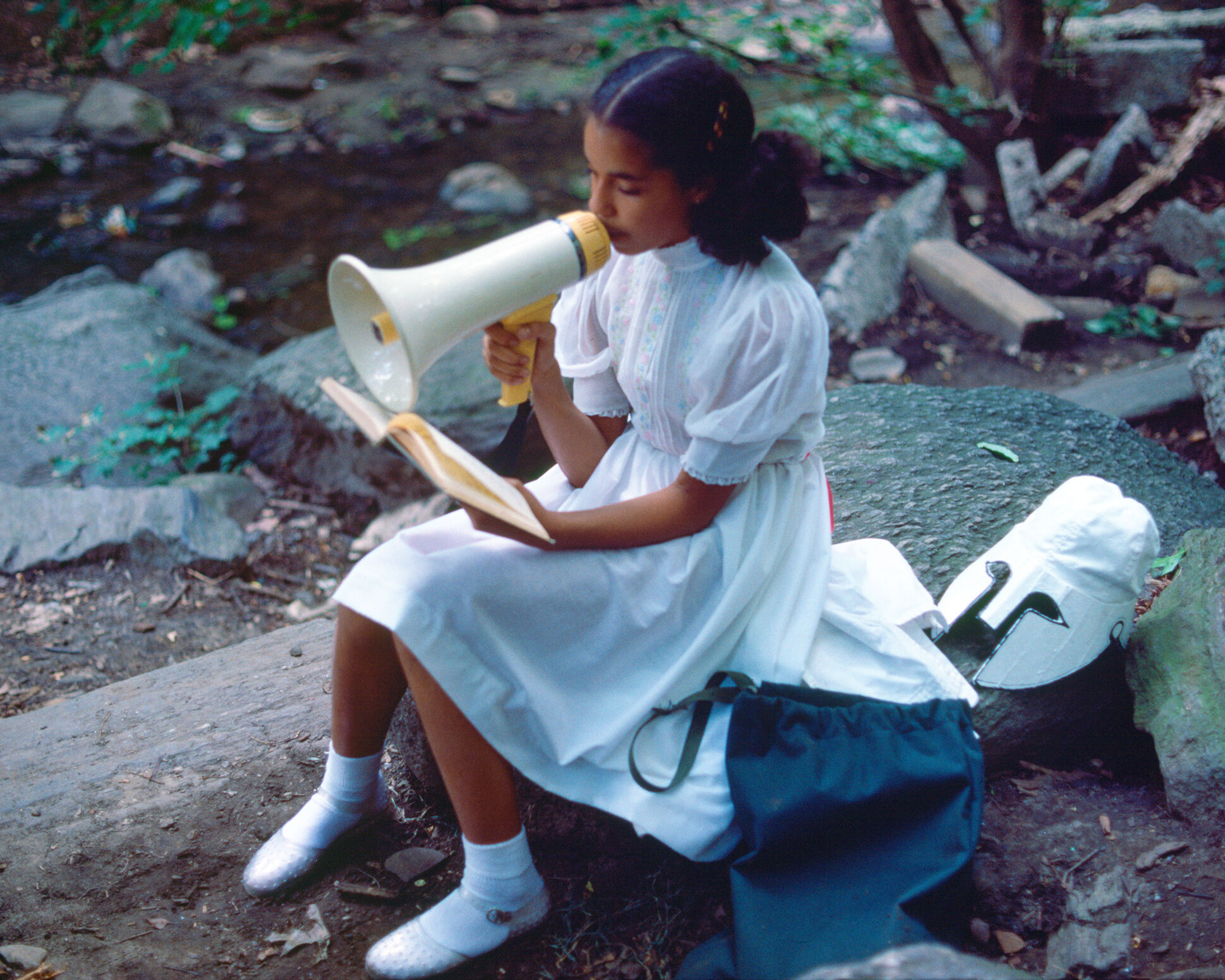 A girl in white holds a microphone sitting on a rock in the forest. 