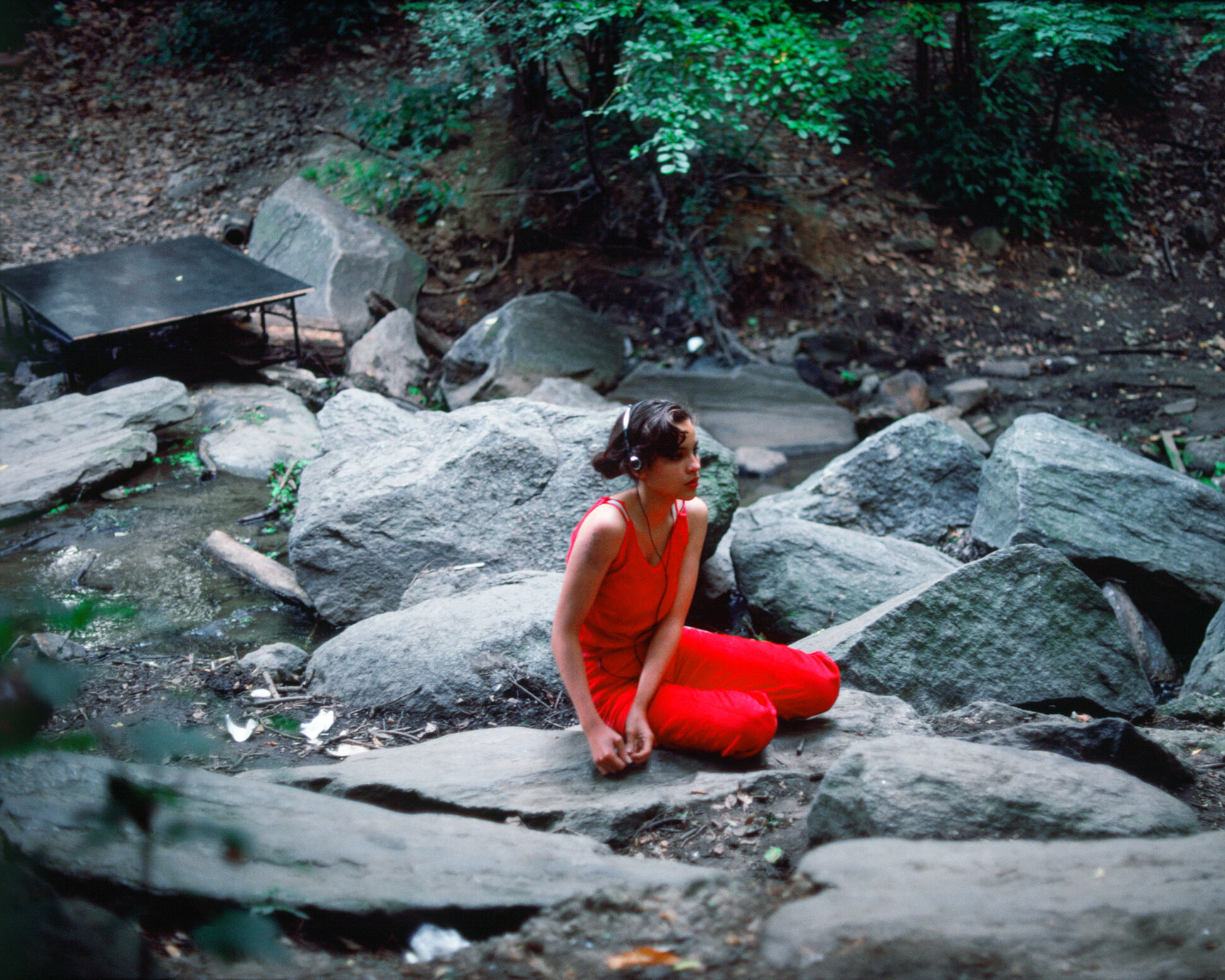 A teenager in magenta sits on a rock in the forest with headphones. 