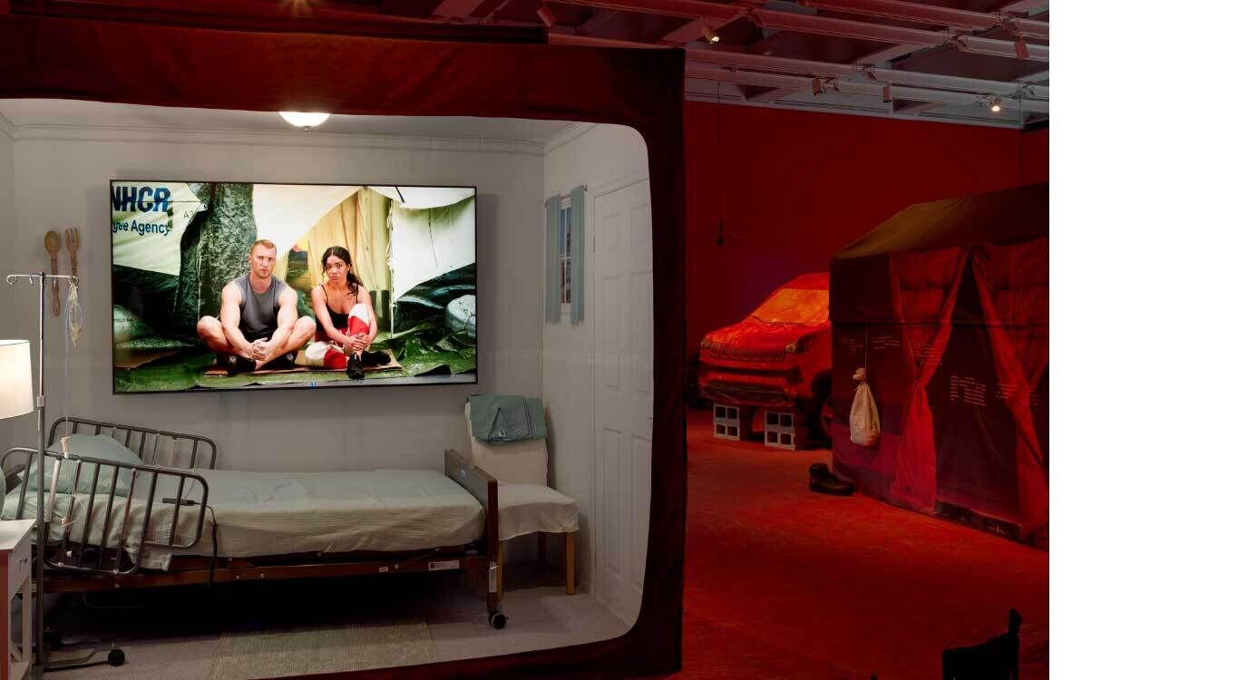 A photograph of a sculptural installation consisting of a temporary shelter for an essential worker in the future when climate change has transformed how we live. The small room contains a hospital bed, a chair, and a bedside table with a lamp on it. Three camp chairs are set-up outside the structure to view the video screen that is mounted within and displays a video interview with workers affected by climate change. The gallery space is illuminated in red light.
