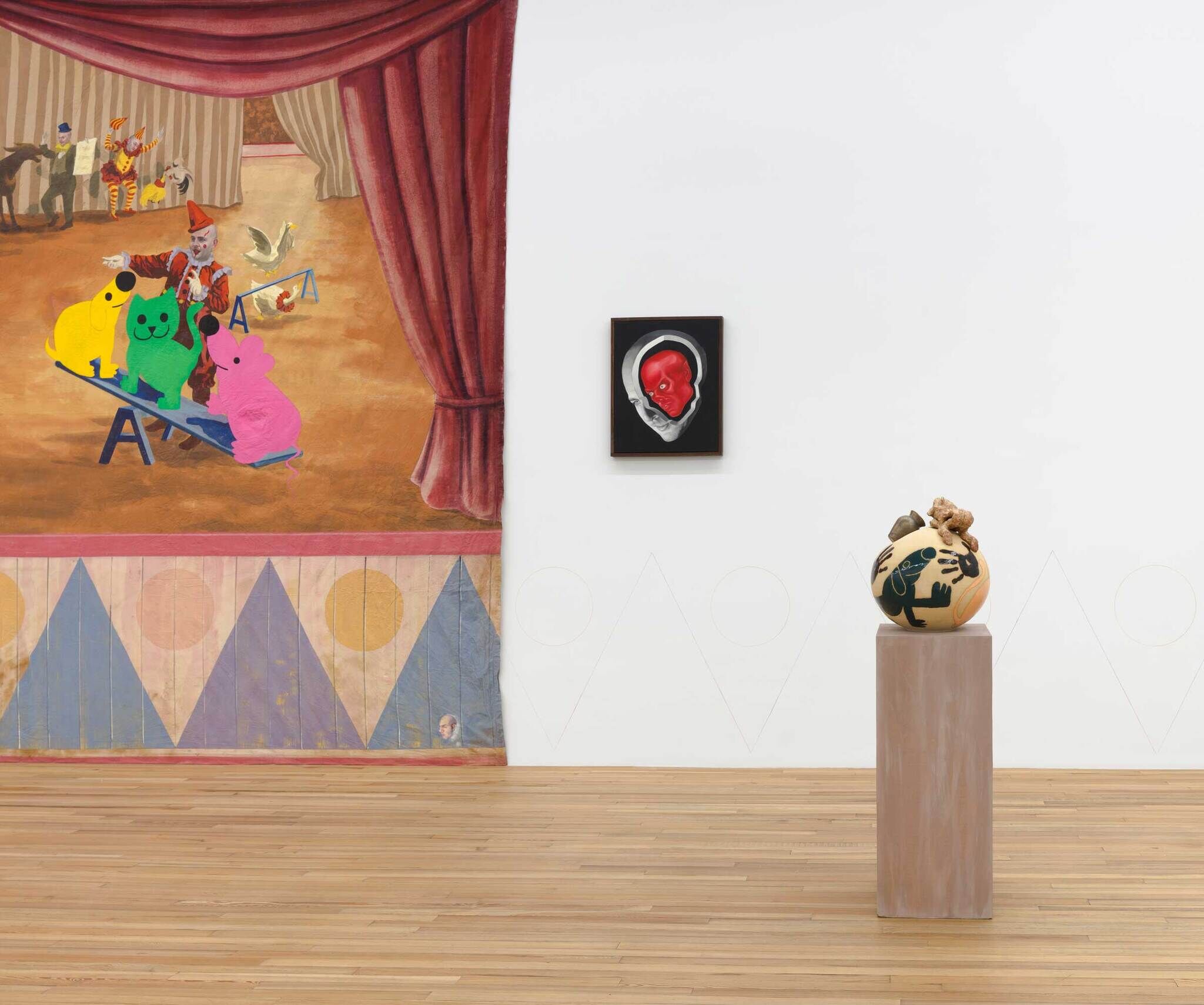 In the background there is a painting of a circus with colorful toy figures next to a smaller painting with a red skull inside of a white skull, and in the foreground there is a circular vase with a black dog.